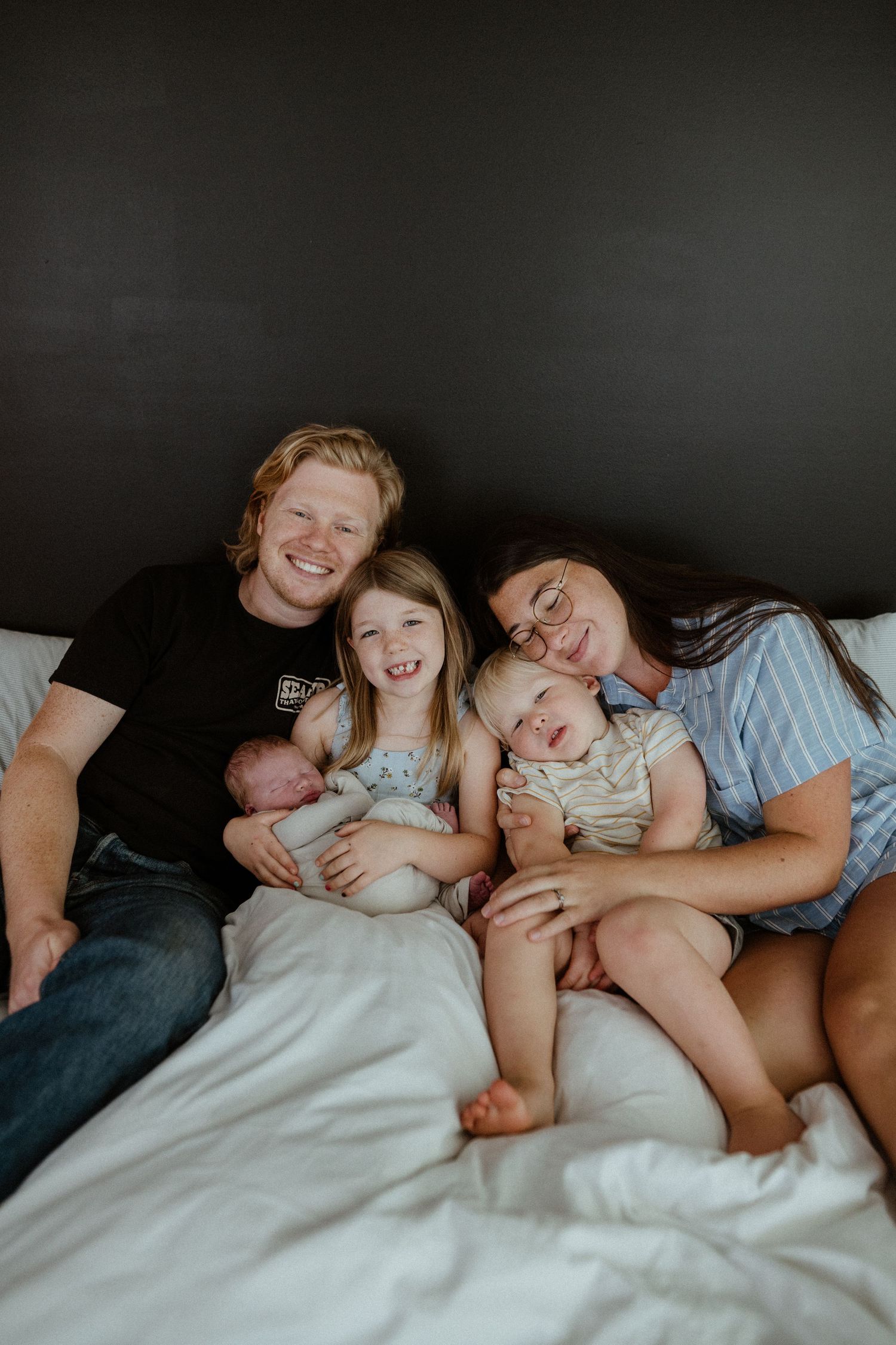 Happy family of four with newborn and toddler smiling together on a white bed.