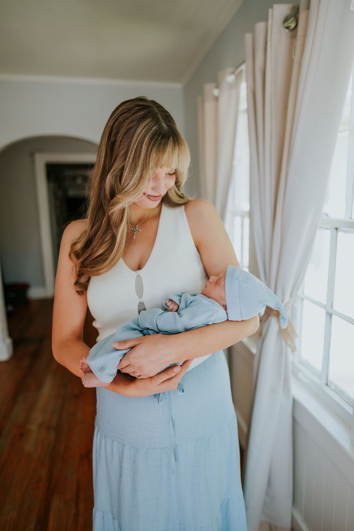 Blonde mother in white top and blue skirt cradling sleeping newborn in blue outfit by bright window.