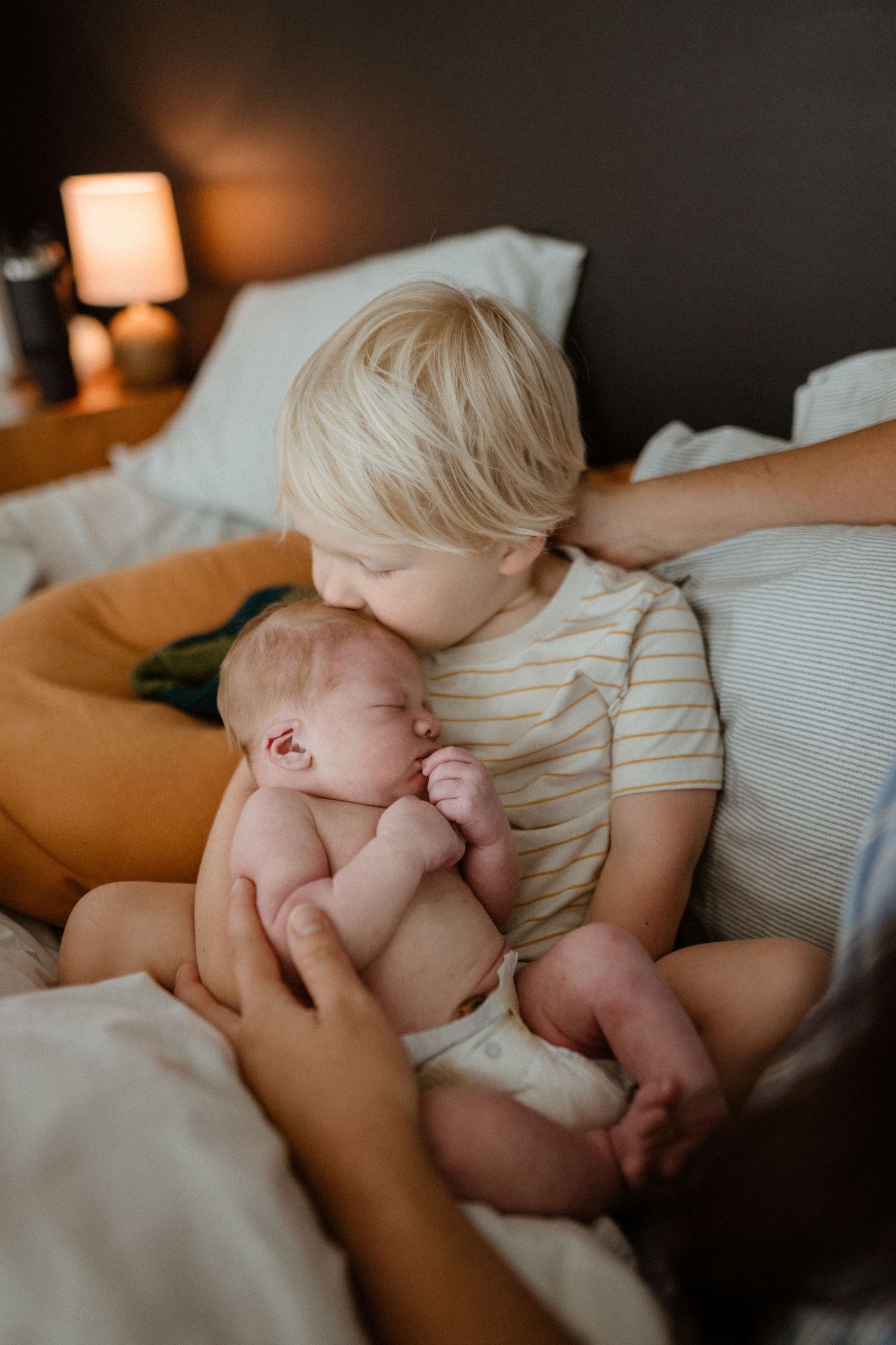 Blonde toddler gently kisses sleeping newborn baby while sitting on white bed with soft lighting.