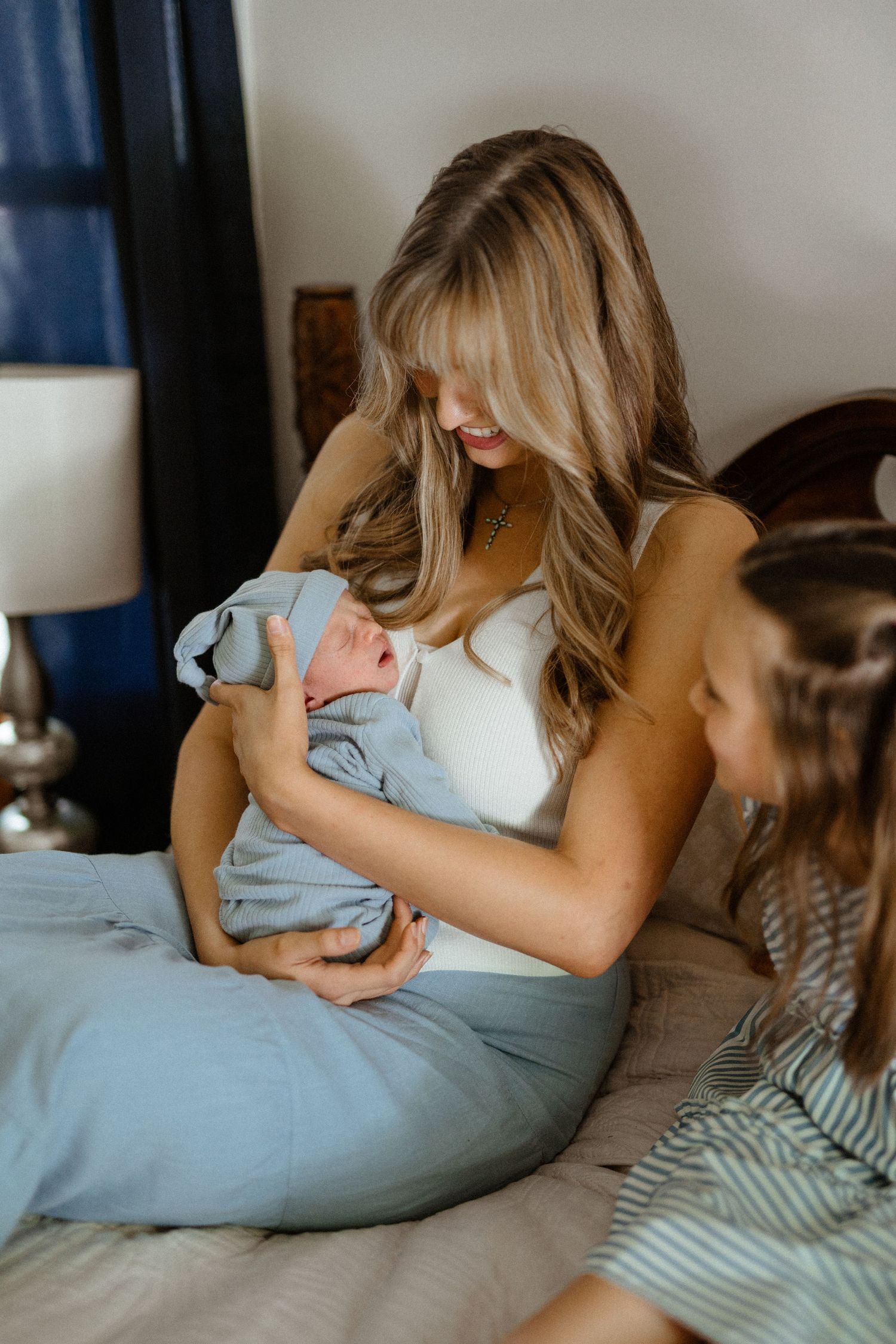 Mother smiling at newborn baby wrapped in blue while older sister looks on during family newborn session.
