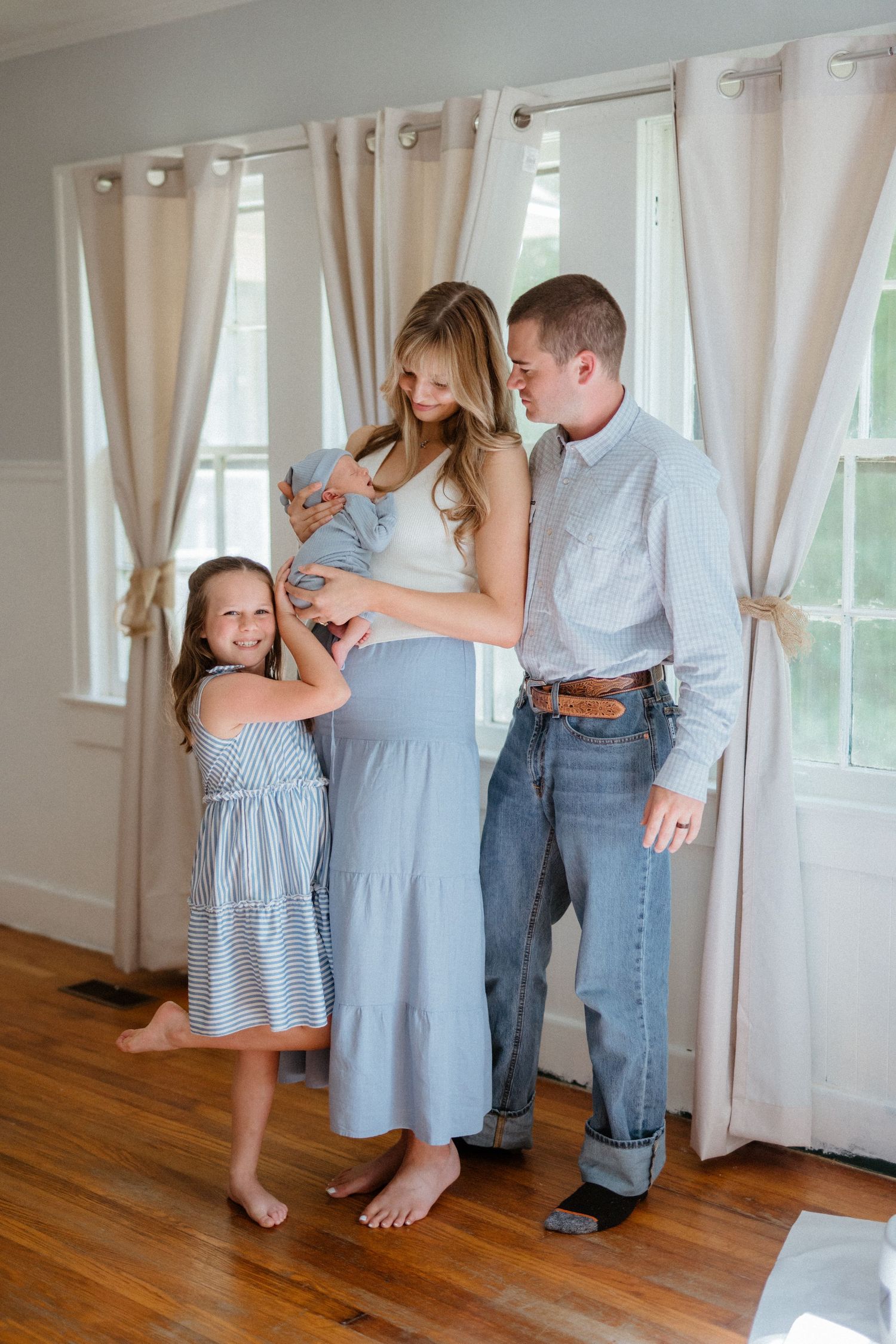 Pregnant woman in blue skirt with husband and daughter touching baby bump indoors.