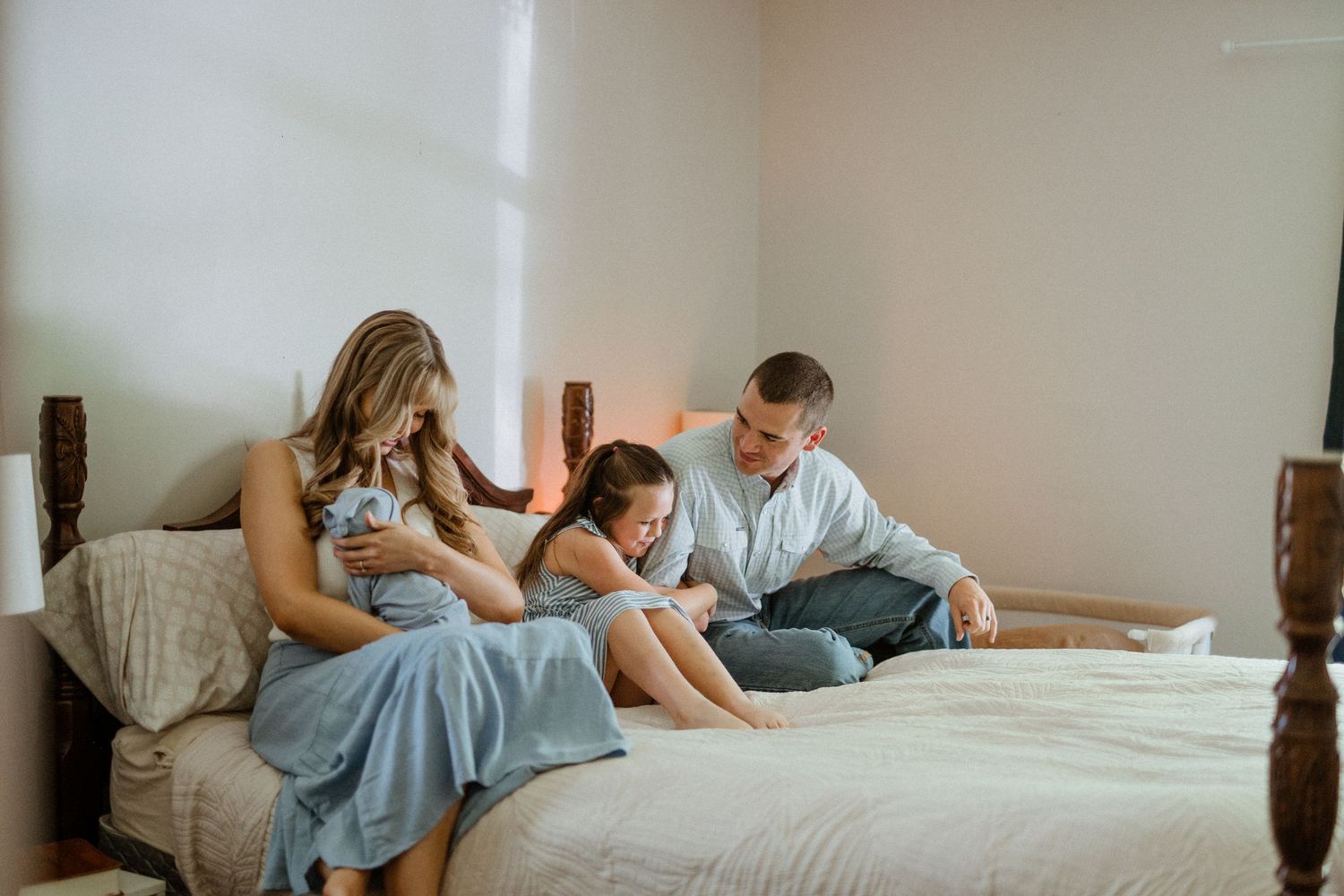 Young family sits together on a bed, parents lovingly attending to their newborn and young daughter.