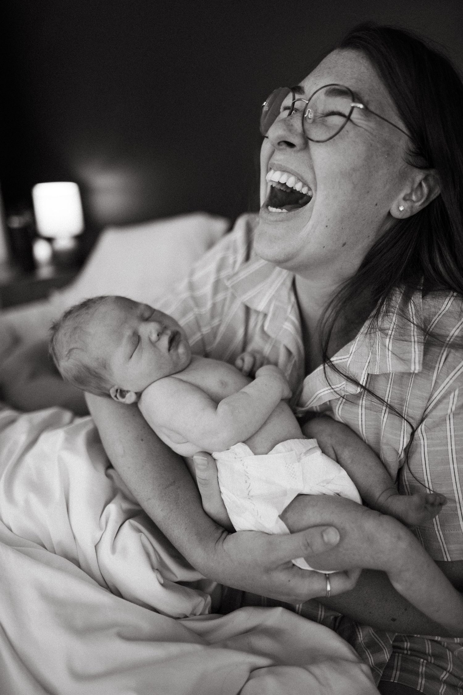 Black and white photo of laughing mother holding newborn baby in bed.