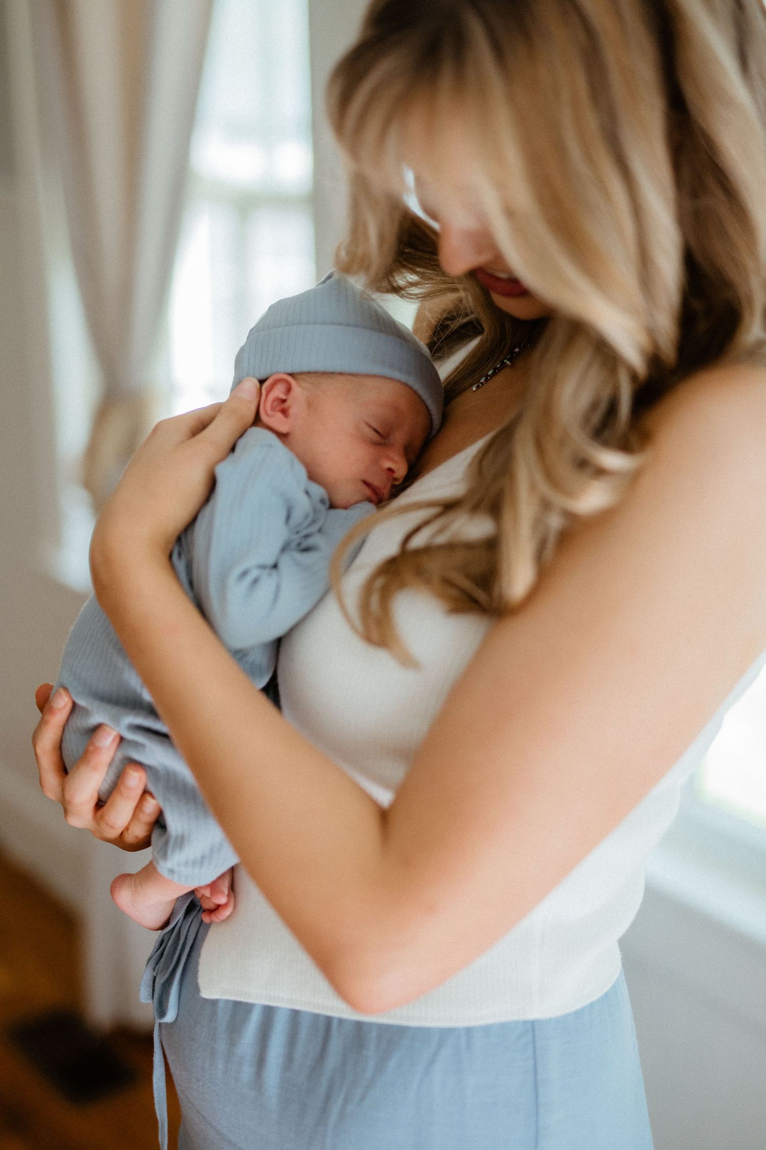 Blonde mother in white tank top gently cradling sleeping newborn in blue outfit near curtained window.