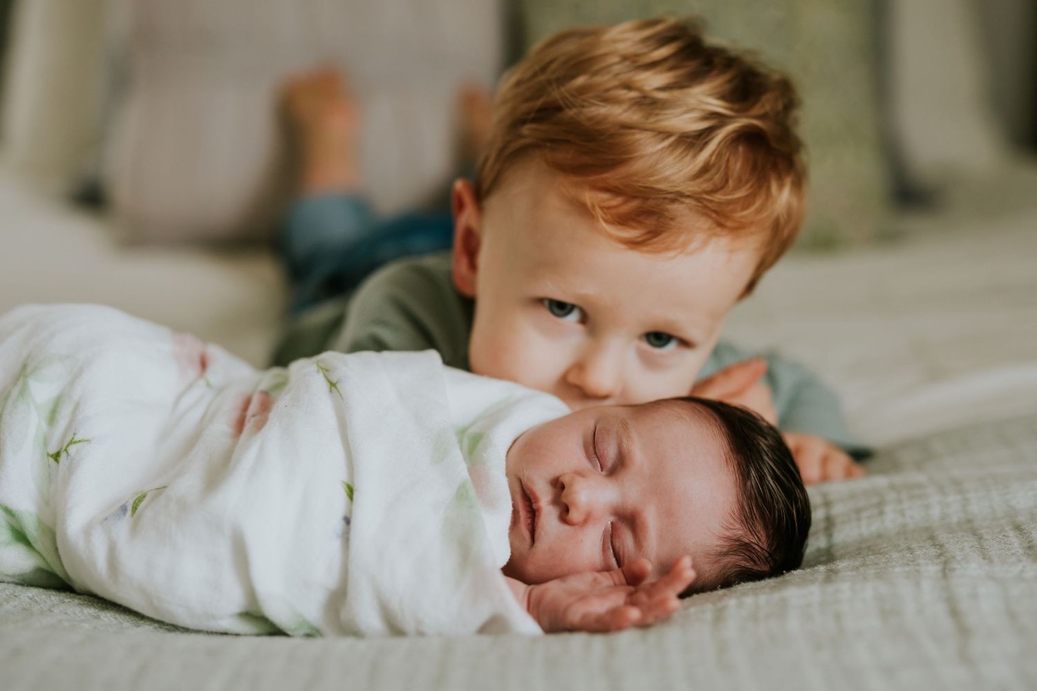 Red-haired toddler boy resting chin on sleeping swaddled newborn baby on a bed.
