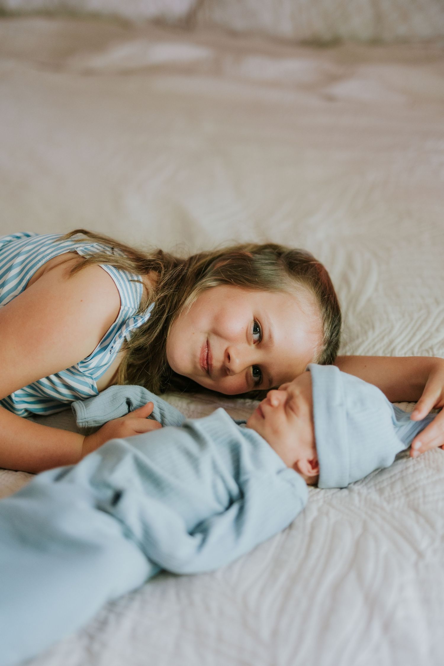 Young girl lying on bed smiling beside newborn baby wrapped in blue blanket.