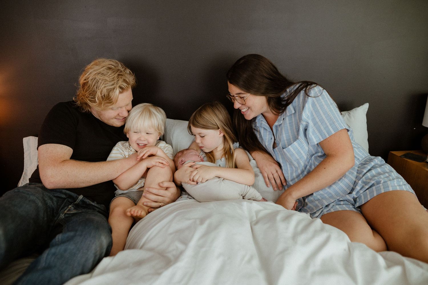 Family of four cuddles on a white bed, parents smiling at newborn baby with two siblings.