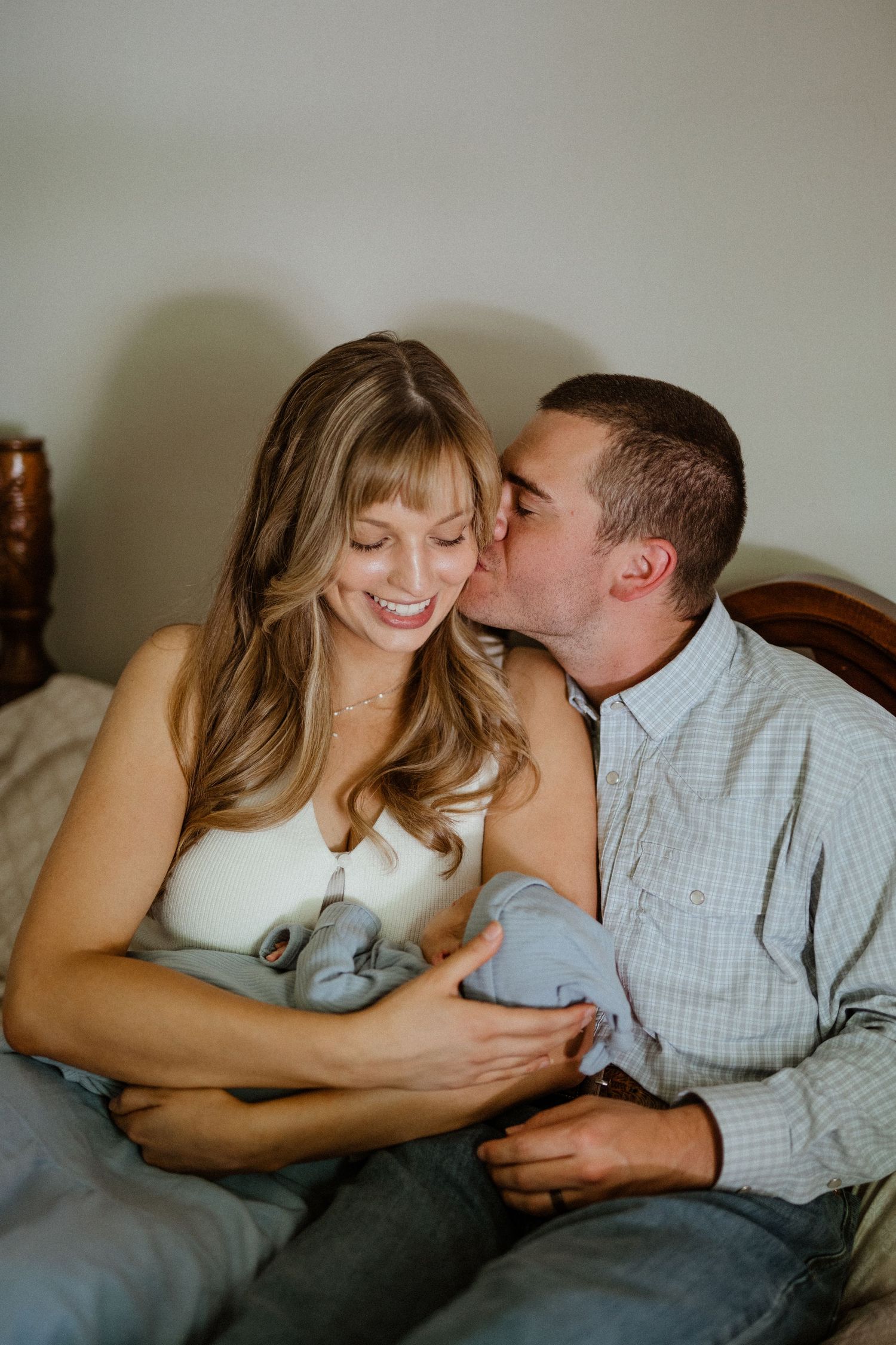 Happy couple sitting on bed with newborn baby as father kisses smiling mother on cheek.