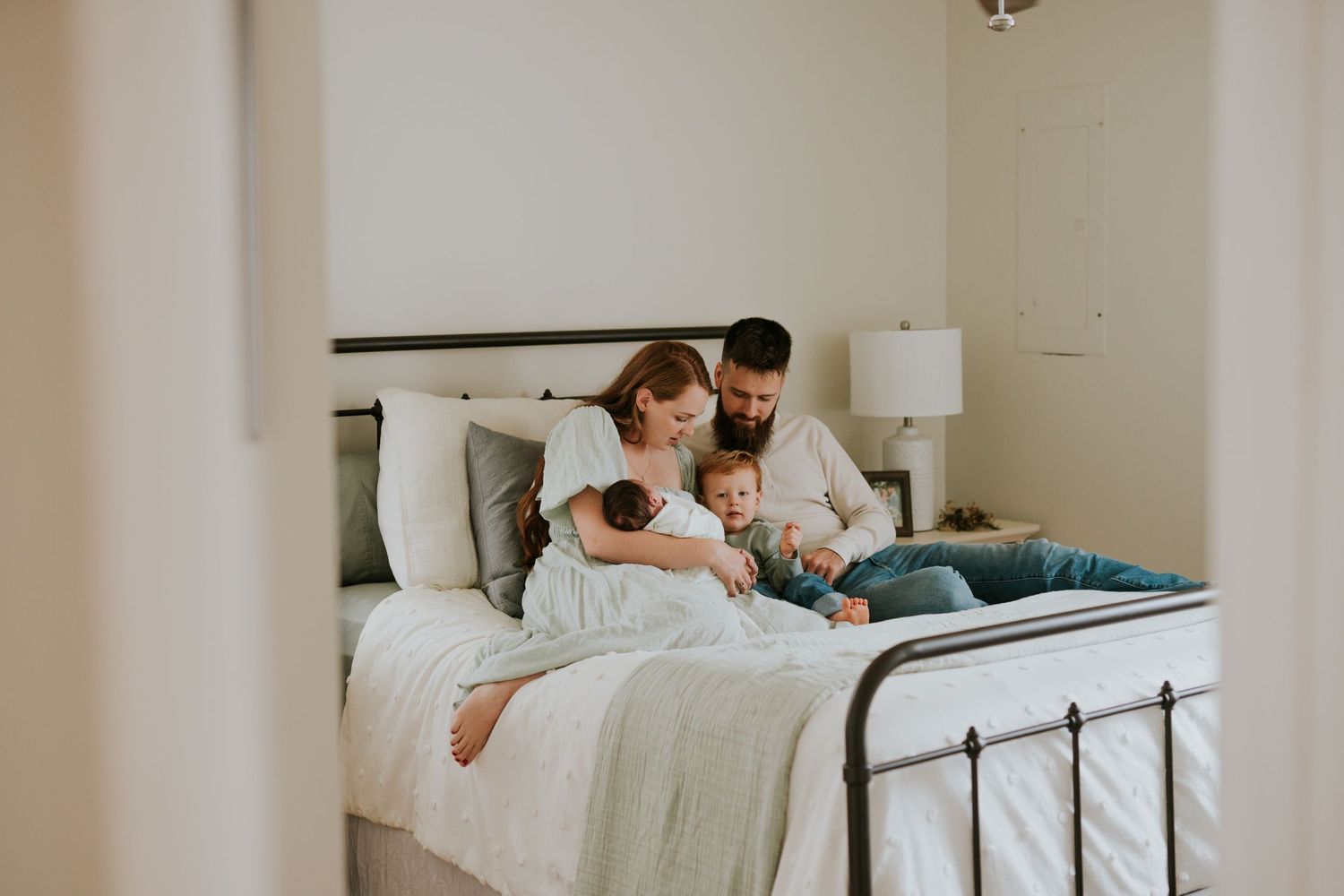 Young family with toddler and newborn cuddling together on a white bed in a bedroom.