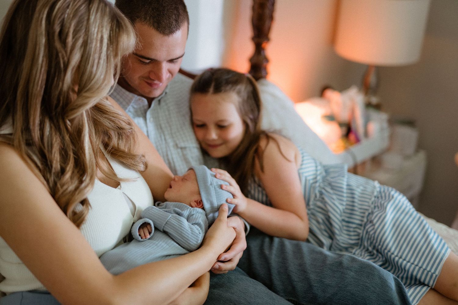 Parents and young daughter admire their newborn baby together on a cozy bed.