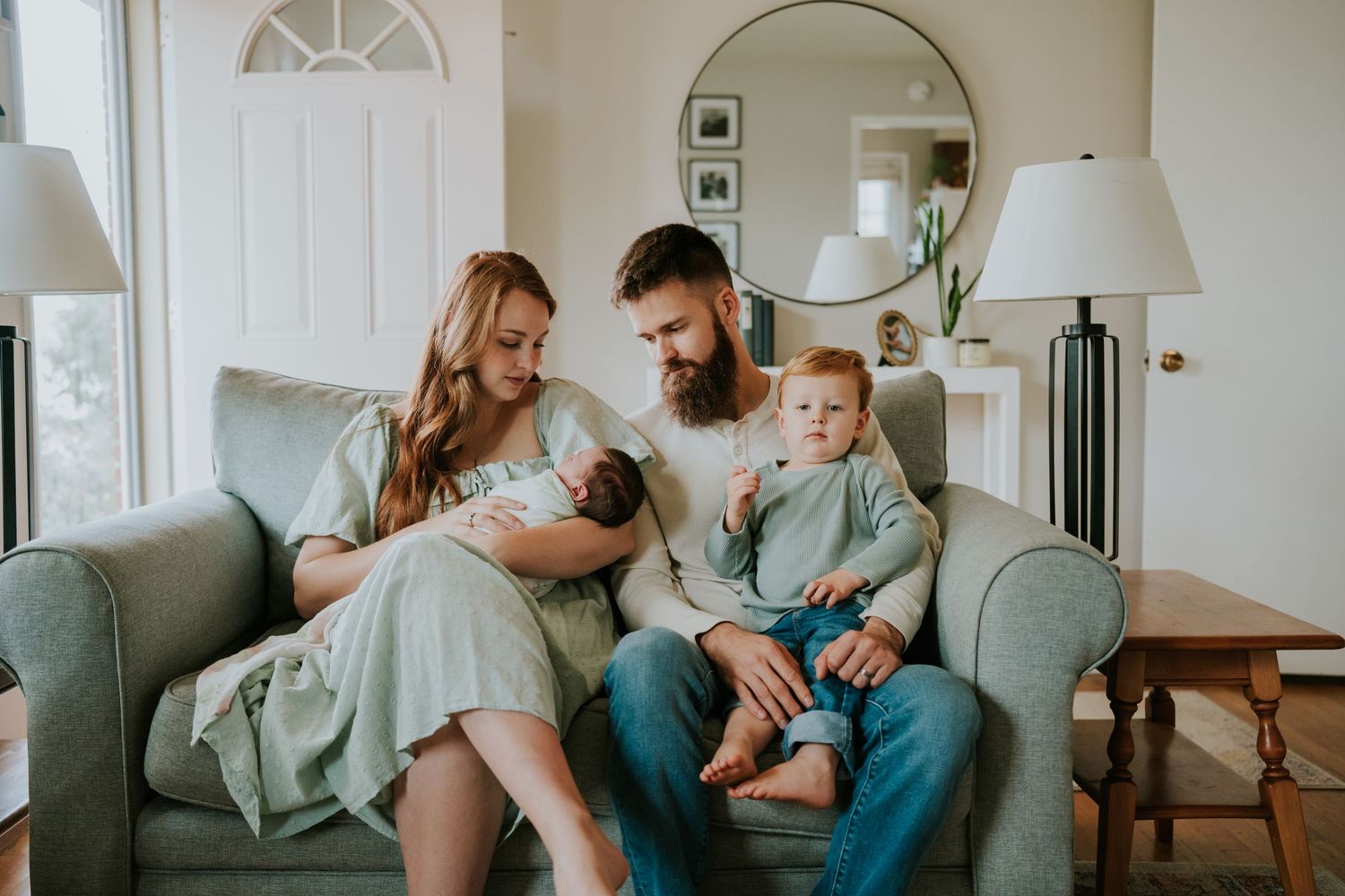 Young family with red-haired toddler and newborn baby sitting together on a gray sofa at home.