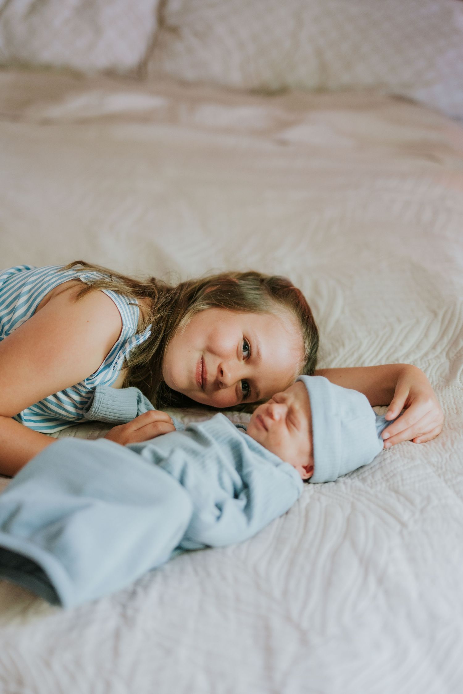 Young girl smiling at camera while lying beside swaddled newborn baby on white bed.