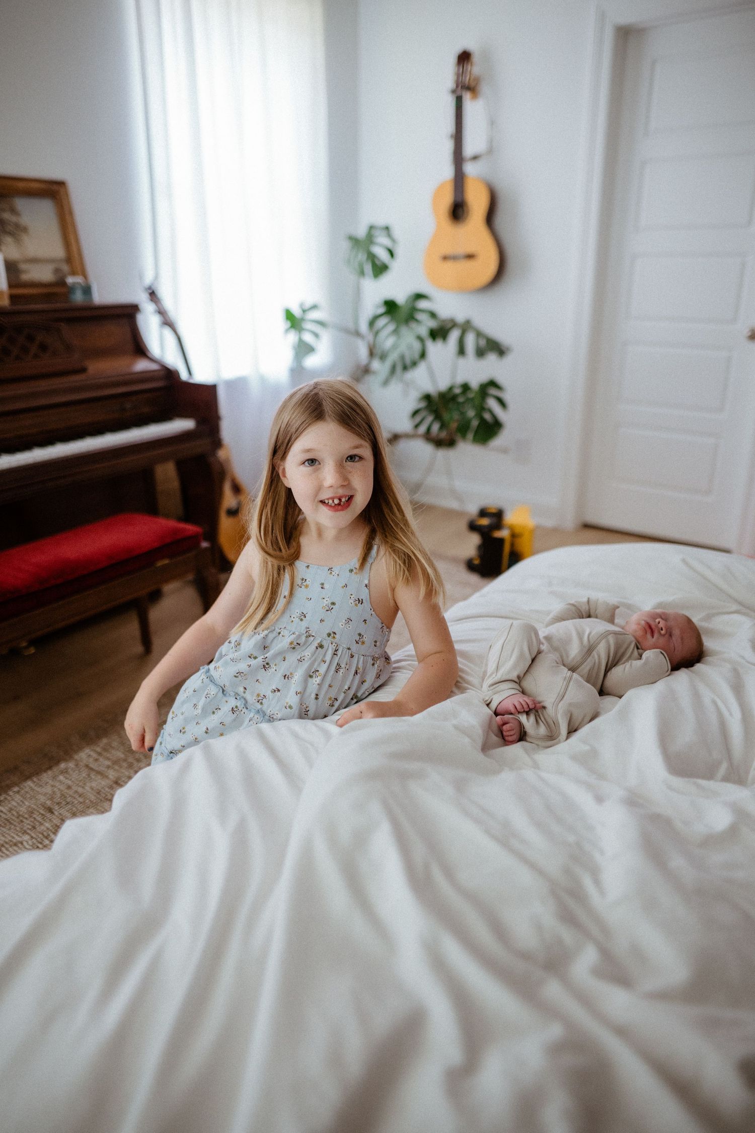Smiling young girl in floral dress sits on white bed with sleeping newborn baby in bright bedroom.
