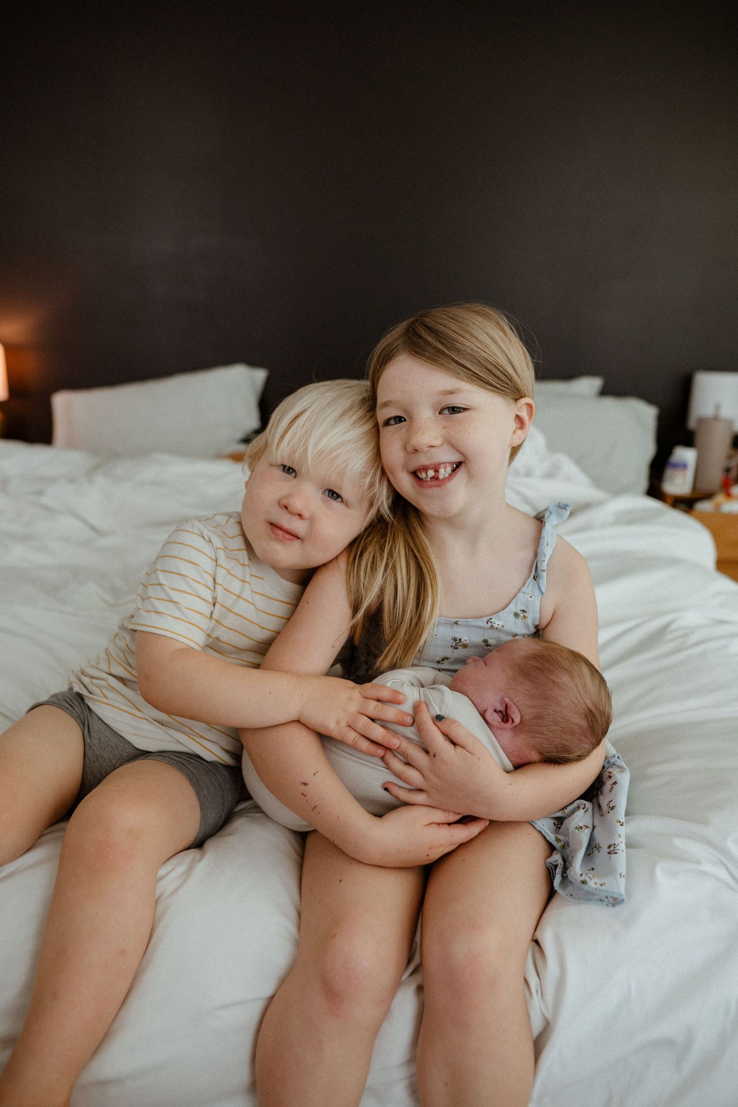 Two young siblings smile on a white bed while the older girl holds a sleeping newborn baby.