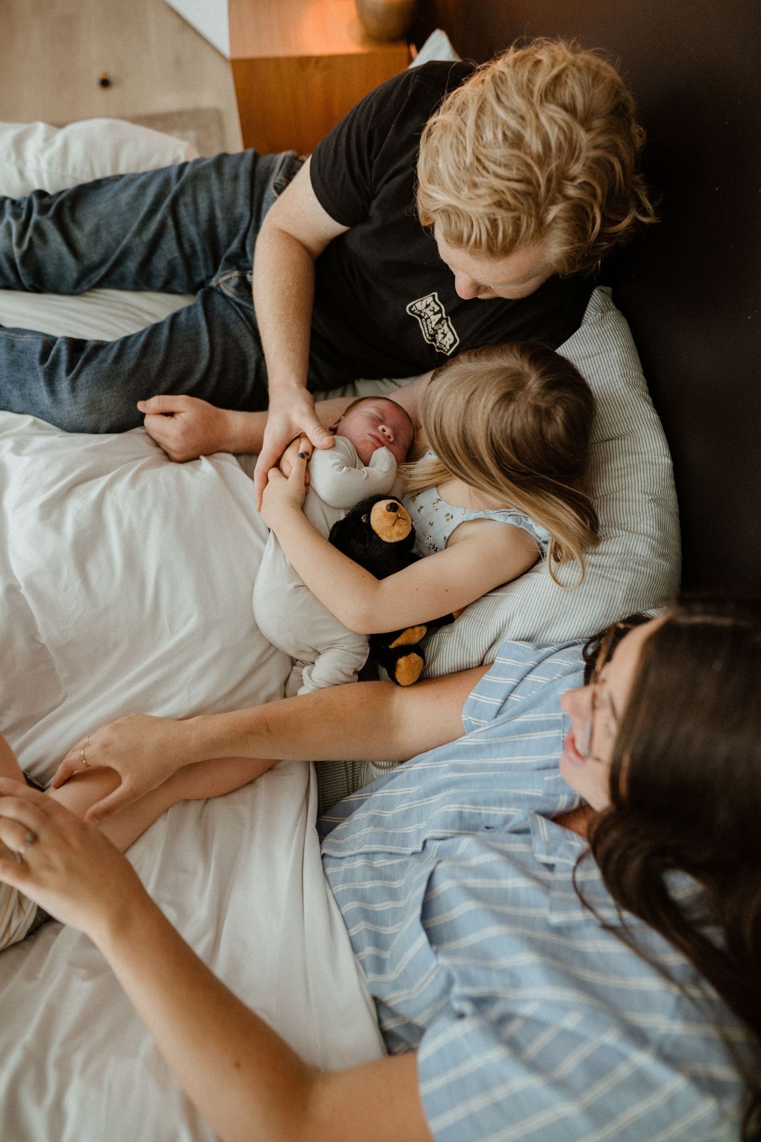 A father and young daughter snuggle together on a bed, lovingly holding a newborn baby.