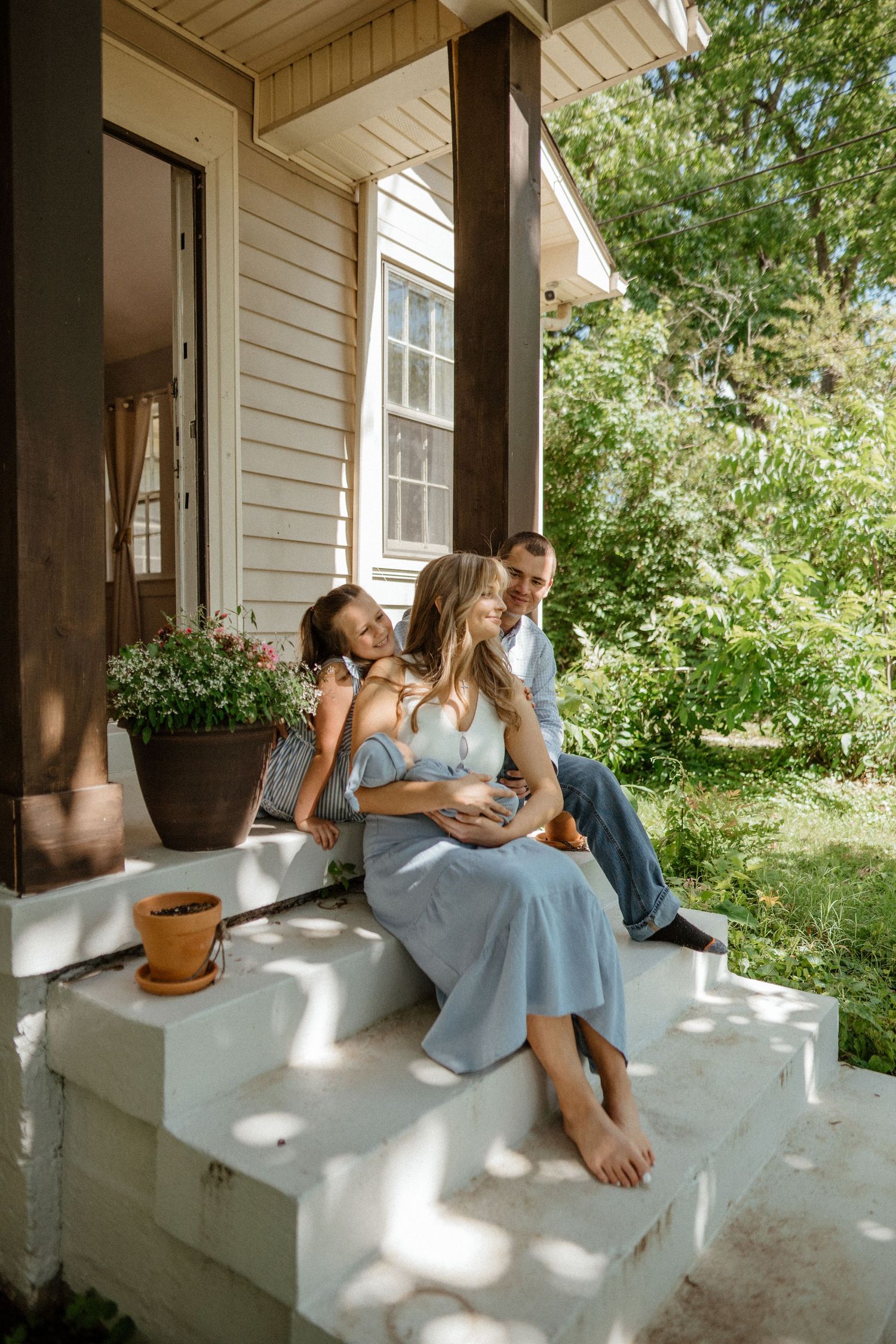 Family of three sits on white porch steps surrounded by lush green garden.