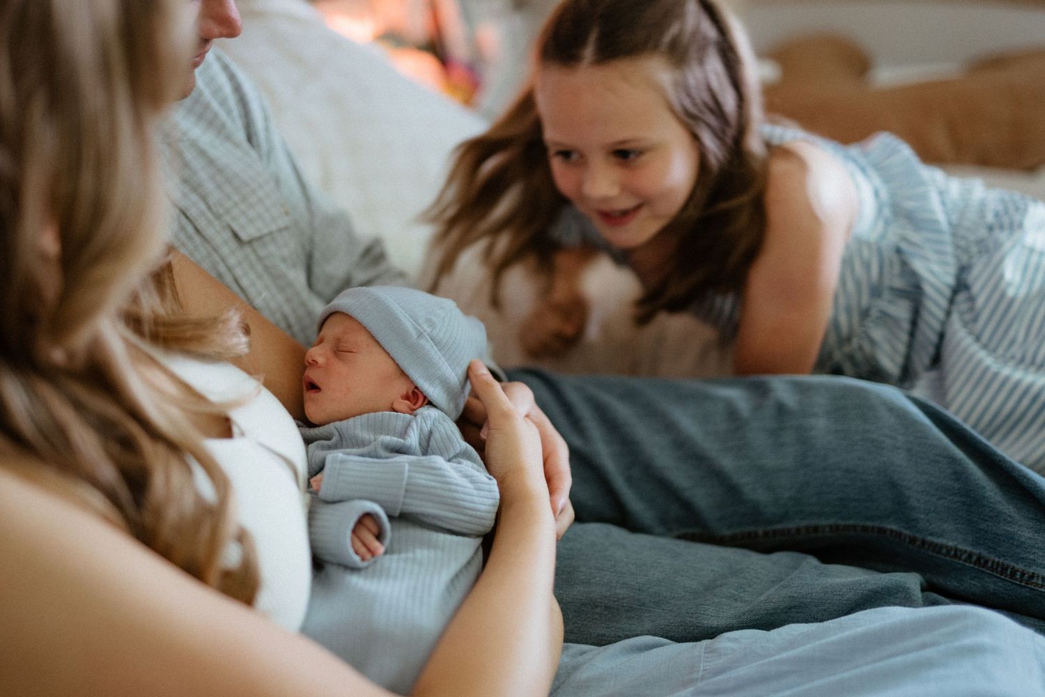 Mother holding newborn baby in blue hat while older sister smiles lovingly nearby.