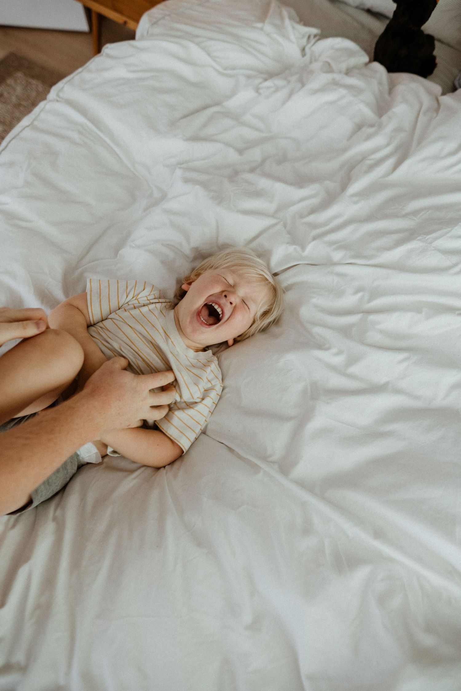 Laughing toddler lies on white bed while adult hands tickle them playfully.