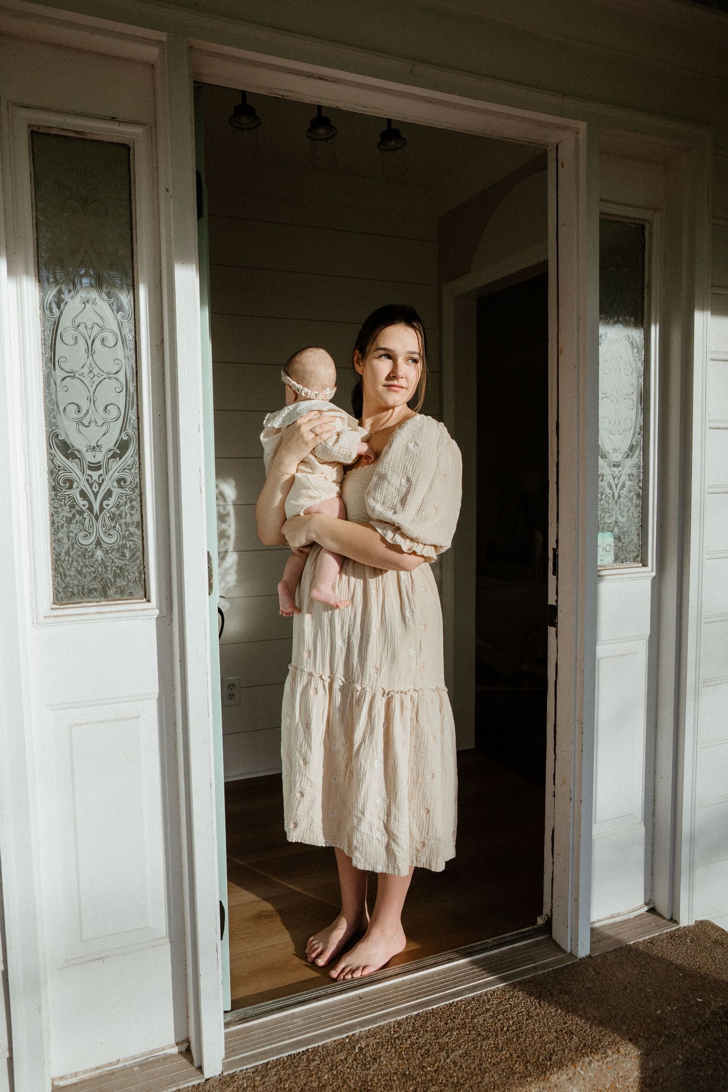 Barefoot mother in cream dress holding newborn baby standing in sunlit doorway.