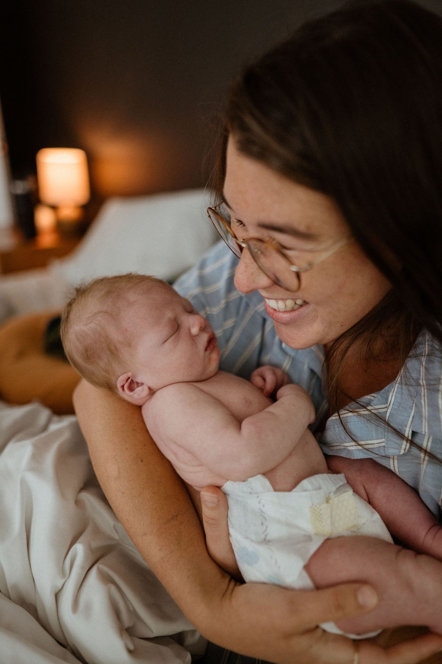 Happy mother with glasses smiles lovingly at her sleeping newborn baby held in her arms.