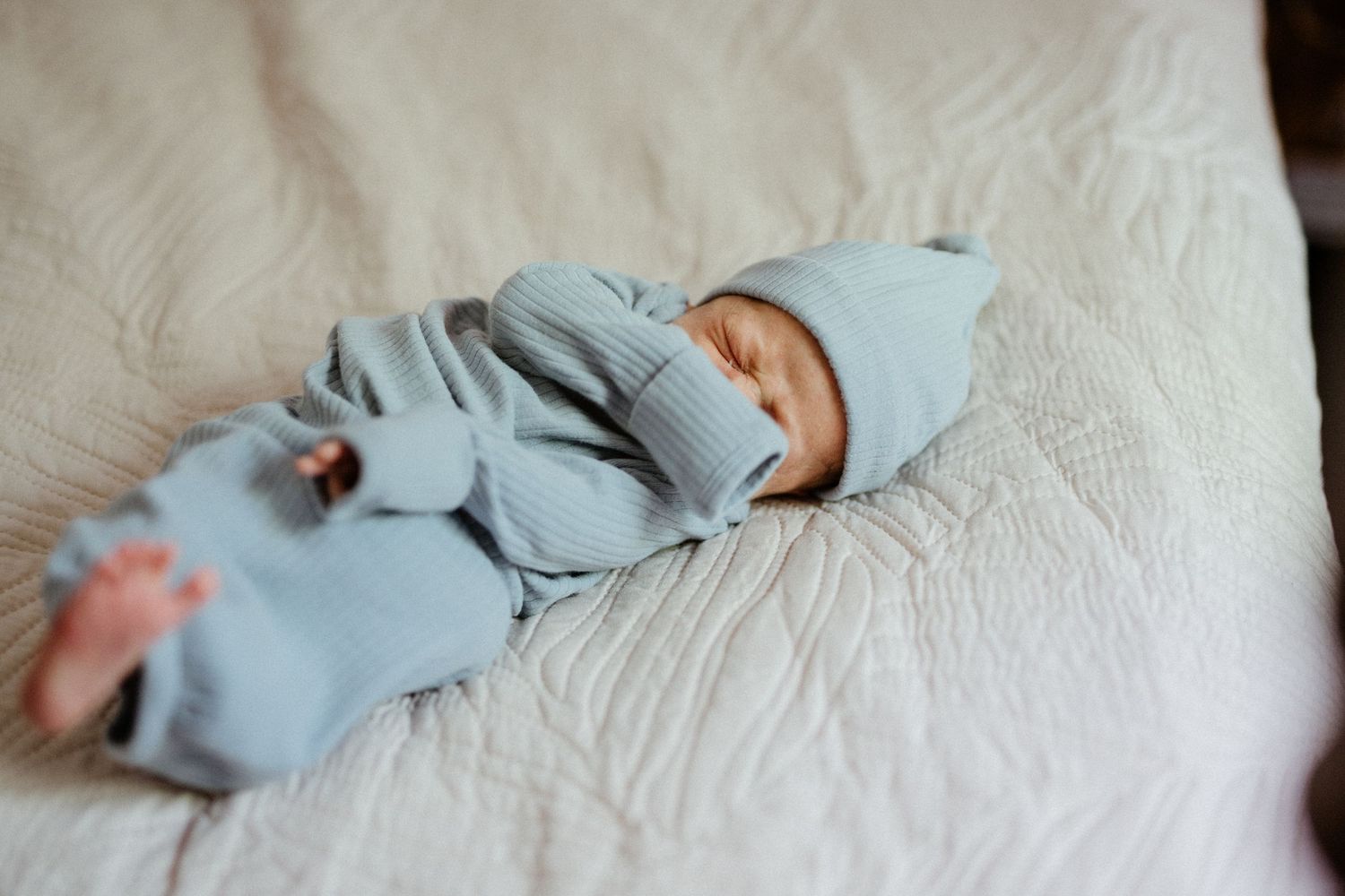 Sleeping newborn baby in light blue knit outfit and hat lying on white quilted bedding.