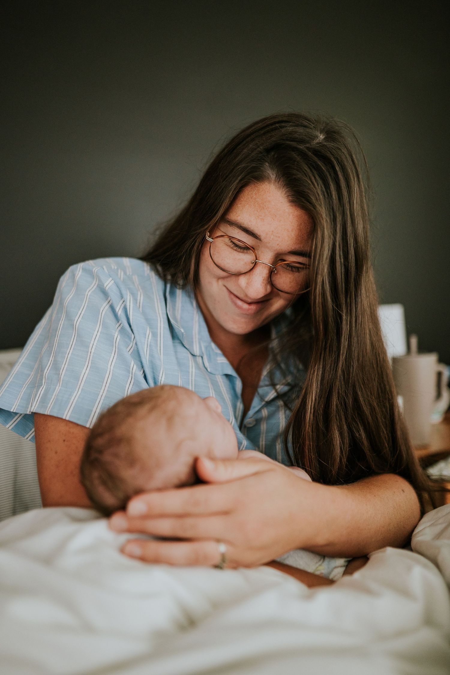 Happy mother with glasses leans over sleeping newborn baby on white bedding.