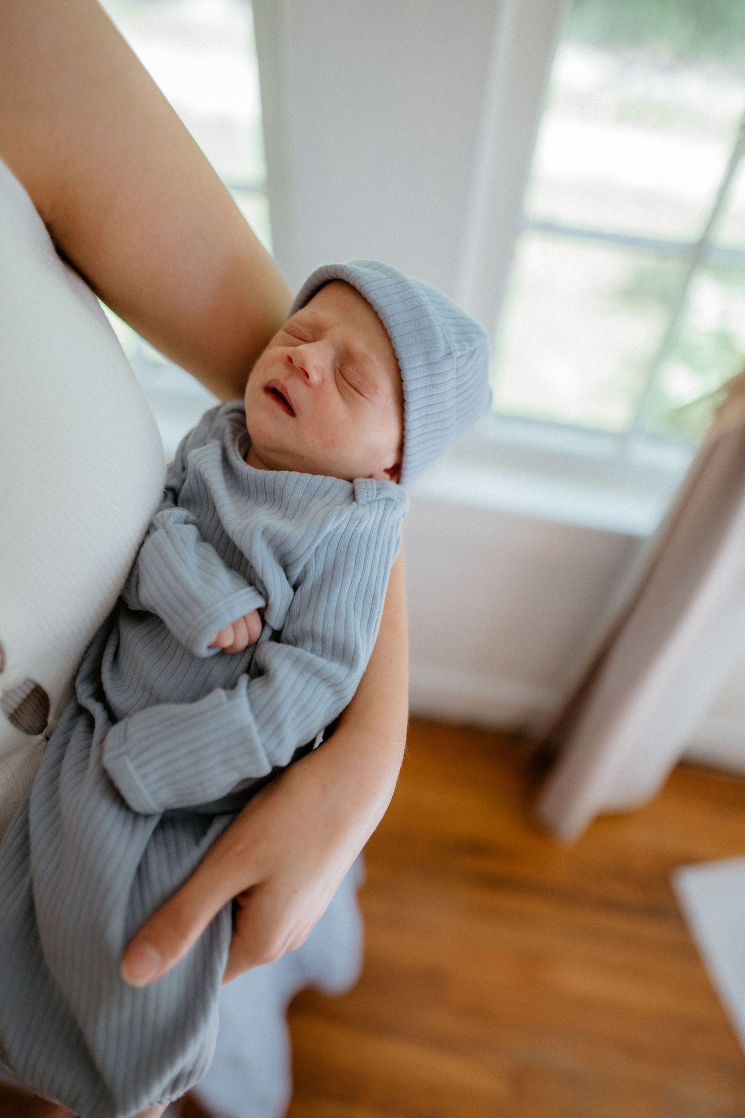 Close-up of newborn baby in blue ribbed outfit and hat being held near sunlit window by mother.