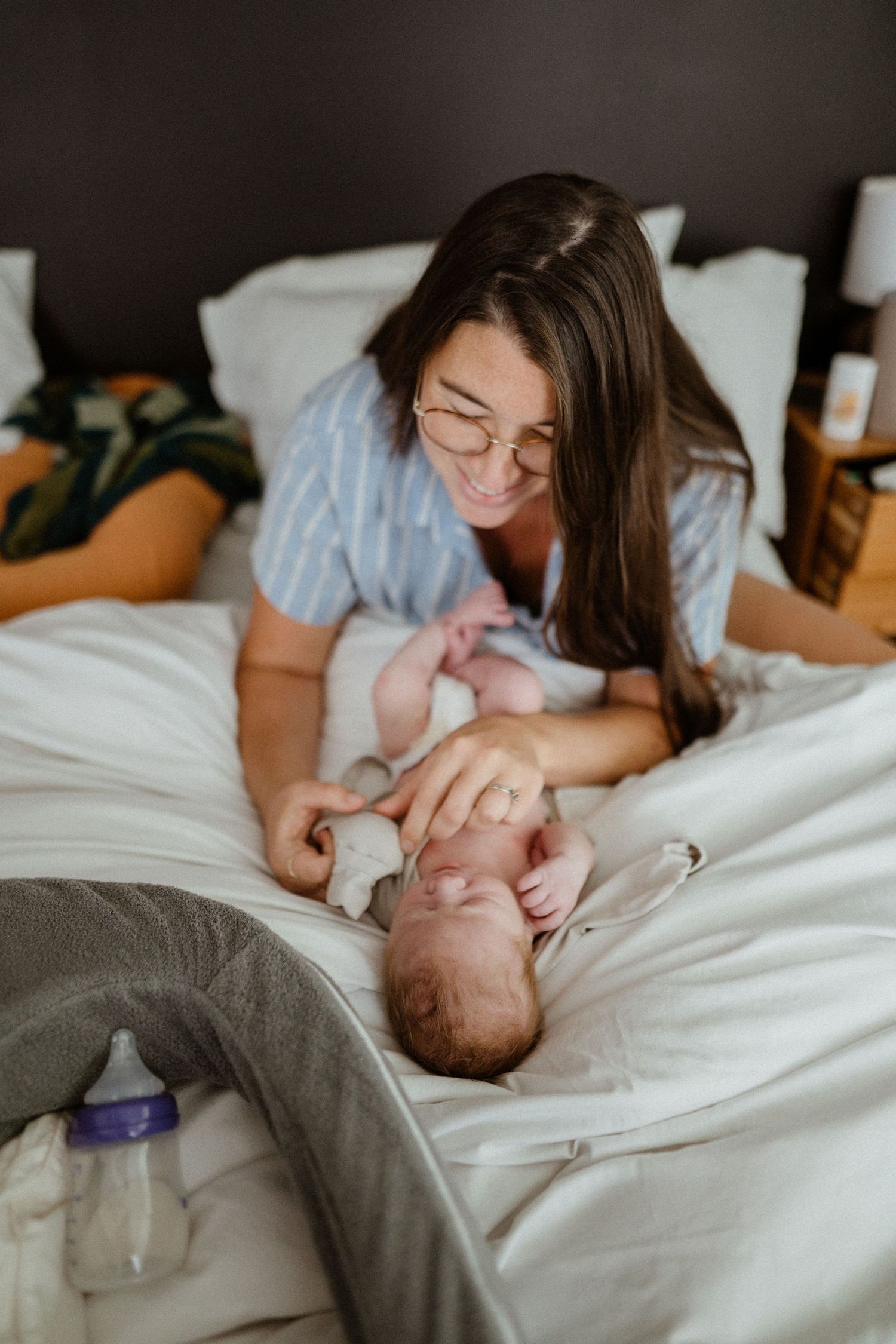 Smiling brunette mother with glasses leans over newborn baby lying on white bed.