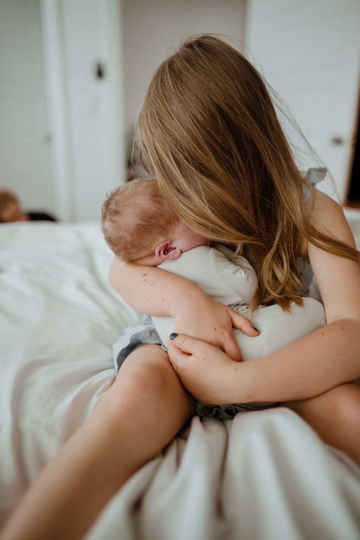 A young girl with blonde hair tenderly holds and cuddles a newborn baby on a white bed.