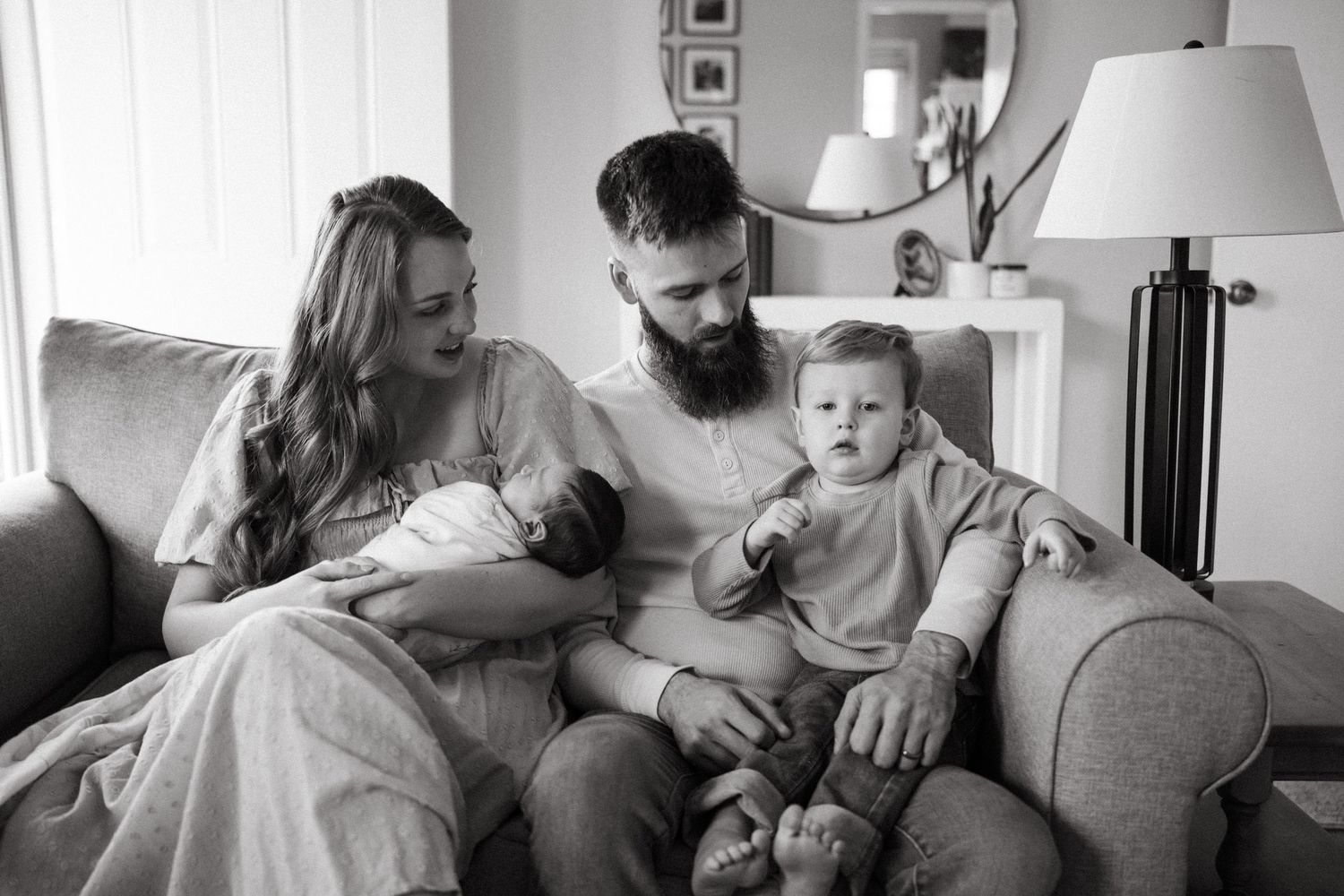 Black and white photo of a young family with a newborn baby and toddler sitting together on a gray sofa.