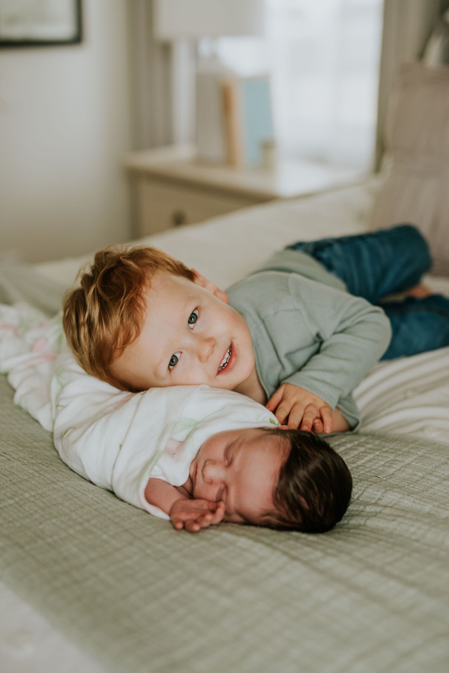 Smiling toddler boy with red hair posing beside sleeping swaddled newborn on a bed.