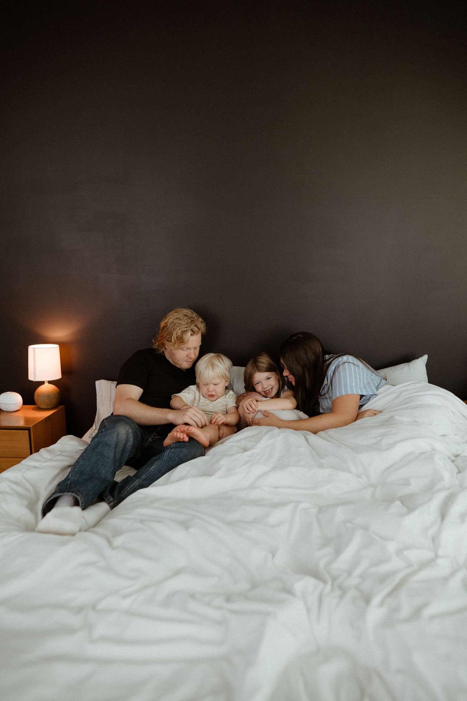 A family of four lies together on a white bed with a dark wall, gazing at their newborn baby.