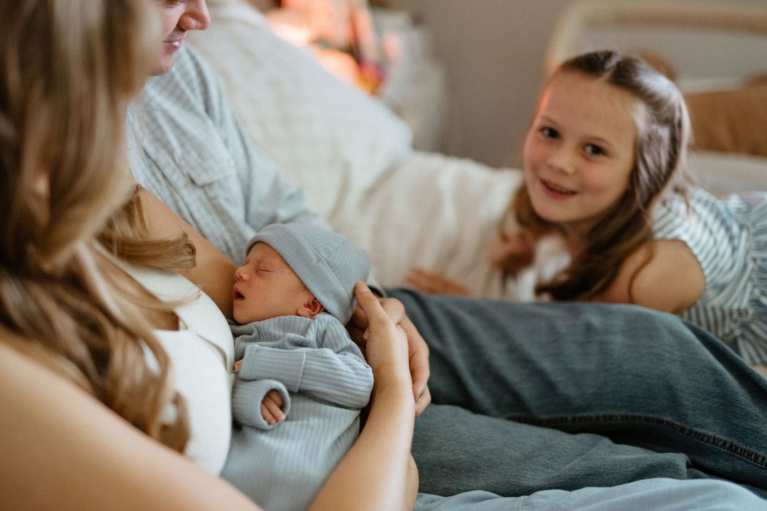 A smiling girl watches her parents tenderly hold their sleeping newborn on a bed.