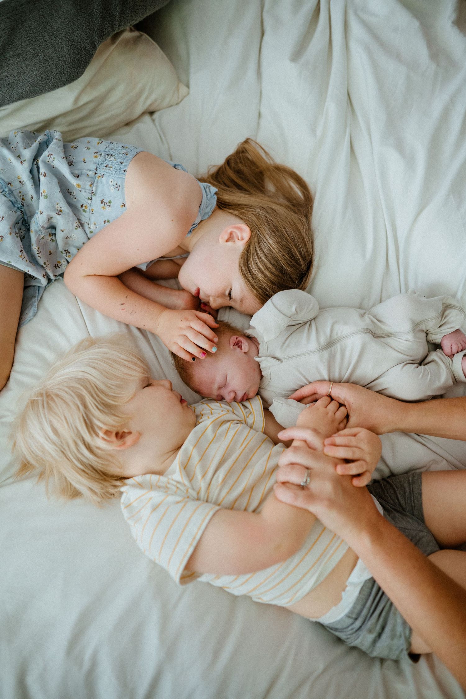 Two young siblings lying on white bed cuddling and kissing their newborn baby brother or sister.