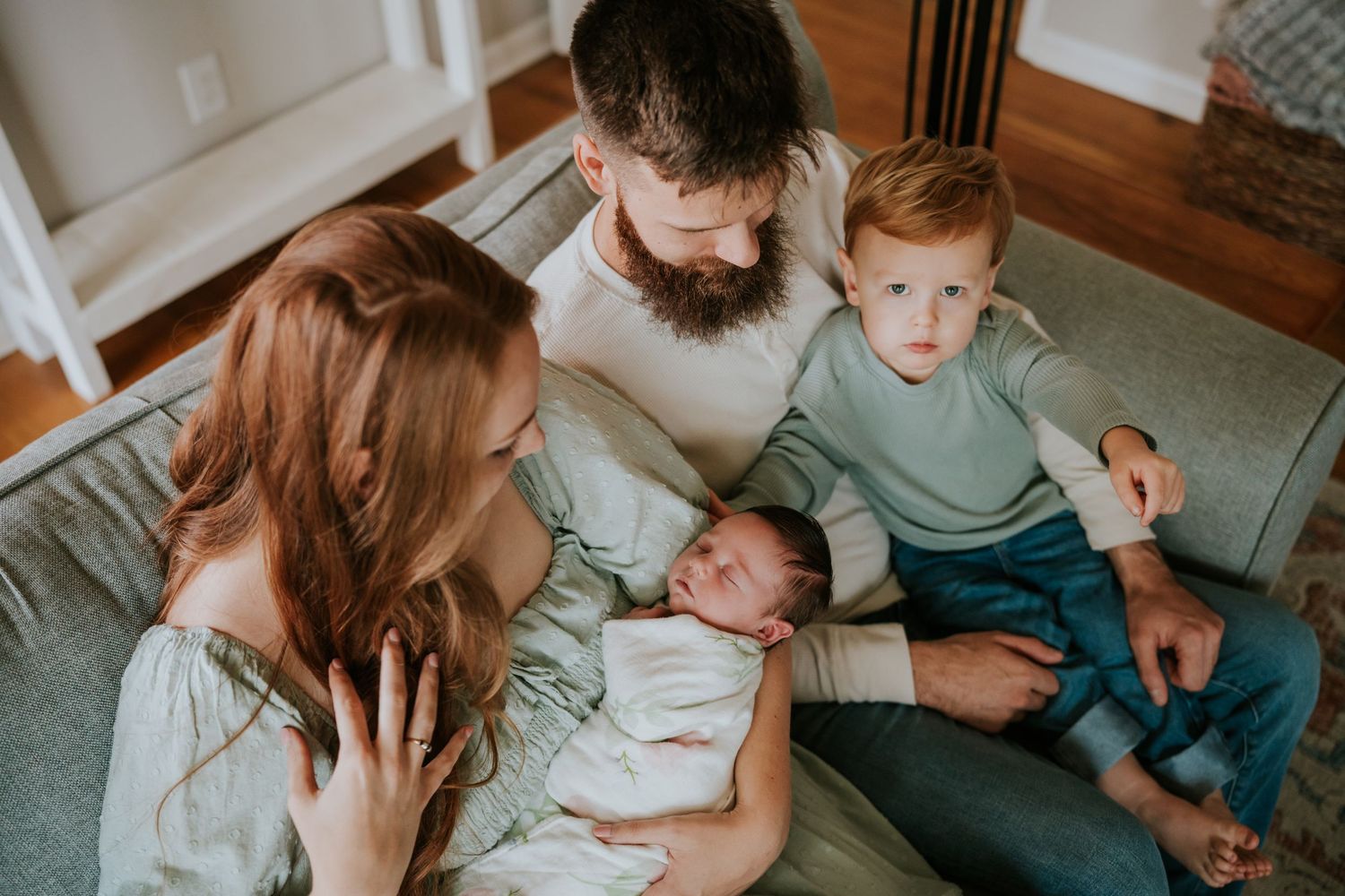 Overhead view of young parents and red-haired toddler admiring their swaddled newborn baby on a sofa.