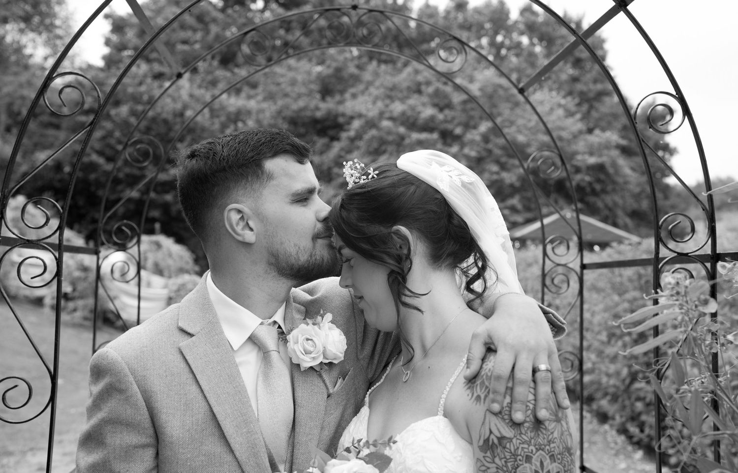 Bride and groom share a romantic kiss under a decorative metal arch in black and white.
