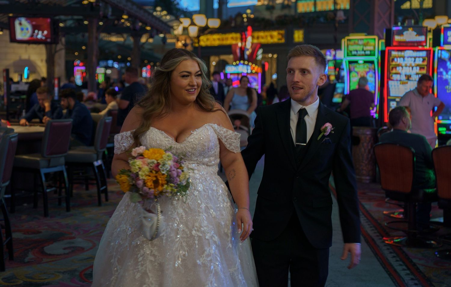 Bride and groom walking through a casino floor, bride holding colorful floral bouquet.