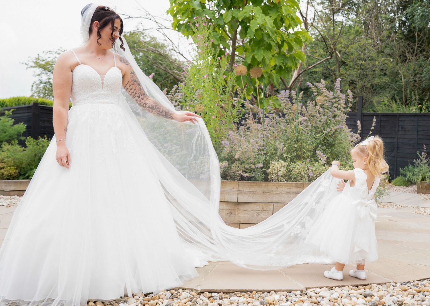 A bride in a white gown and veil stands with a young flower girl in a garden setting.