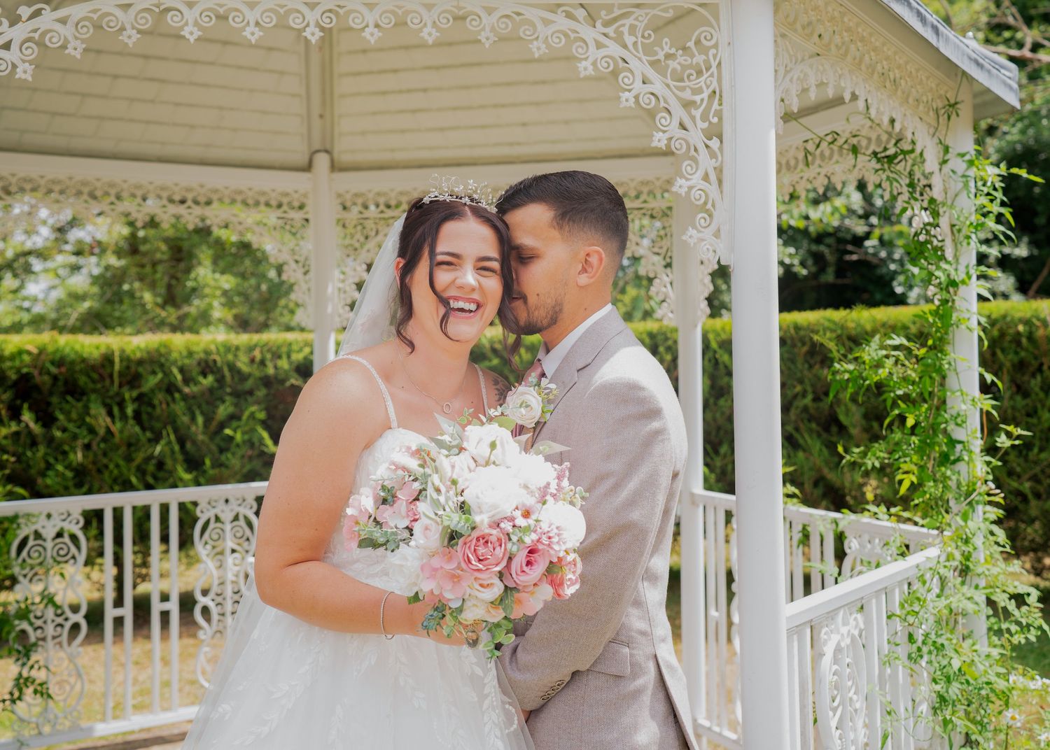 Bride and groom embracing and smiling under a white gazebo surrounded by lush greenery, bride holding pink bouquet.