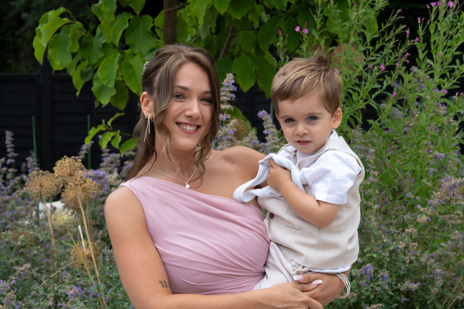 A young woman in a pink dress holds a toddler boy surrounded by lush garden flowers.