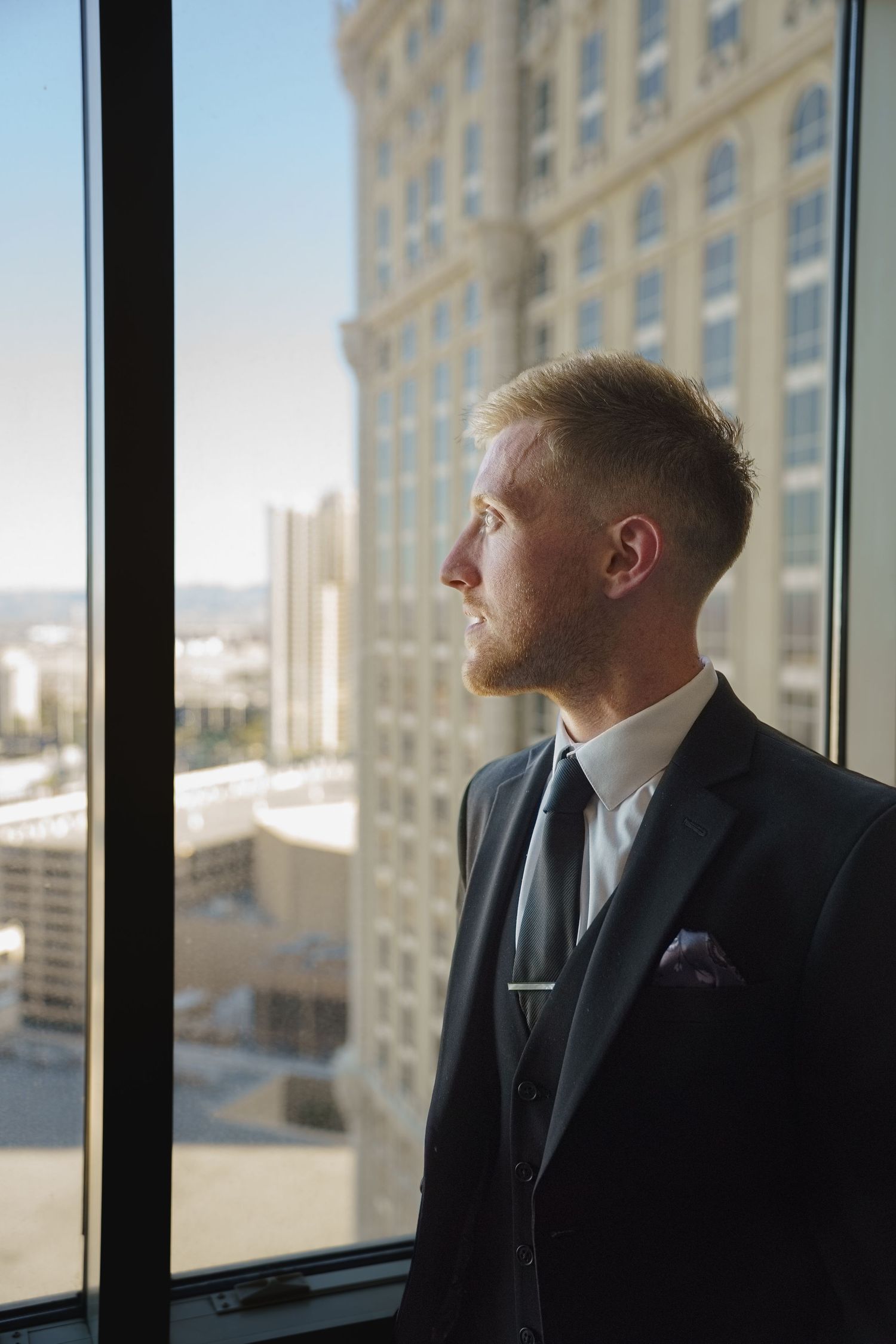 Handsome groom in dark suit gazing out a large window overlooking a Las Vegas cityscape.