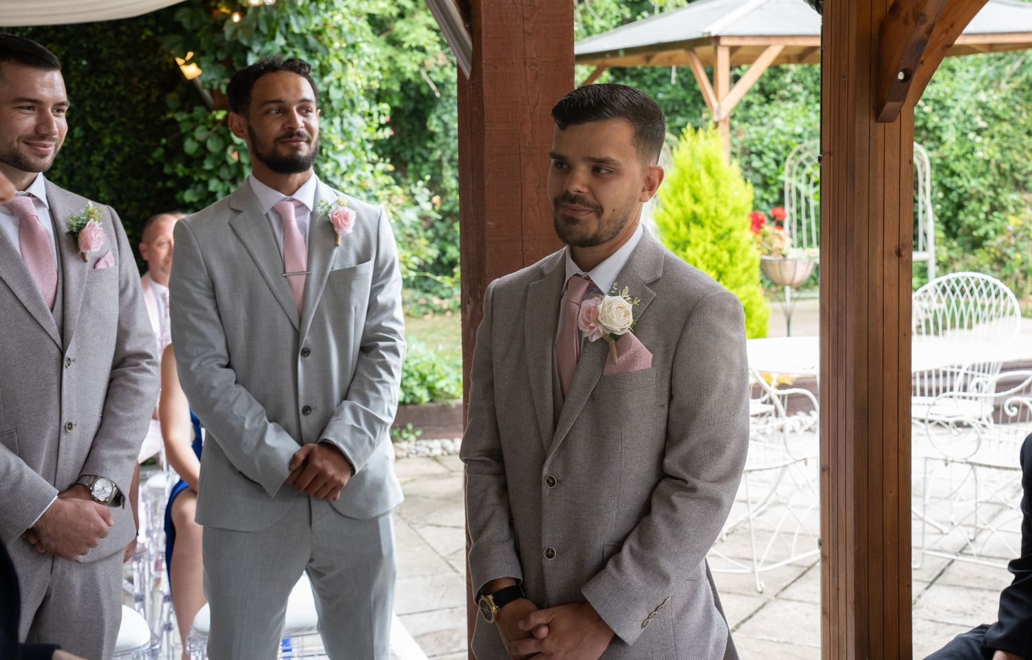 Groom in grey suit watching bride walk down aisle at outdoor wedding ceremony.