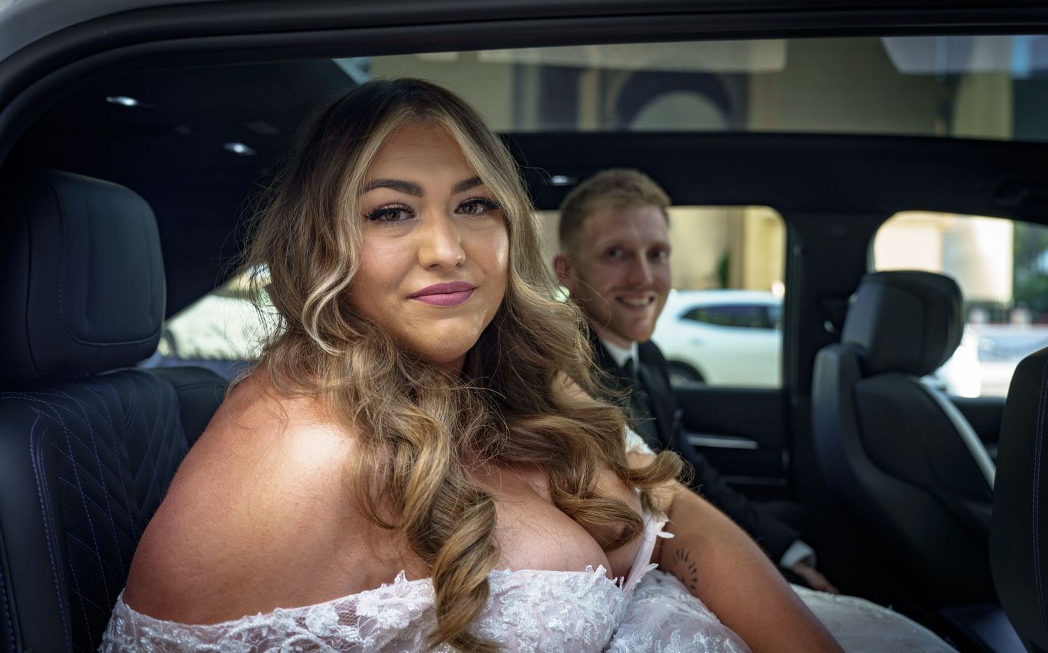 Smiling bride in lace wedding dress sits in limousine with groom visible in background.