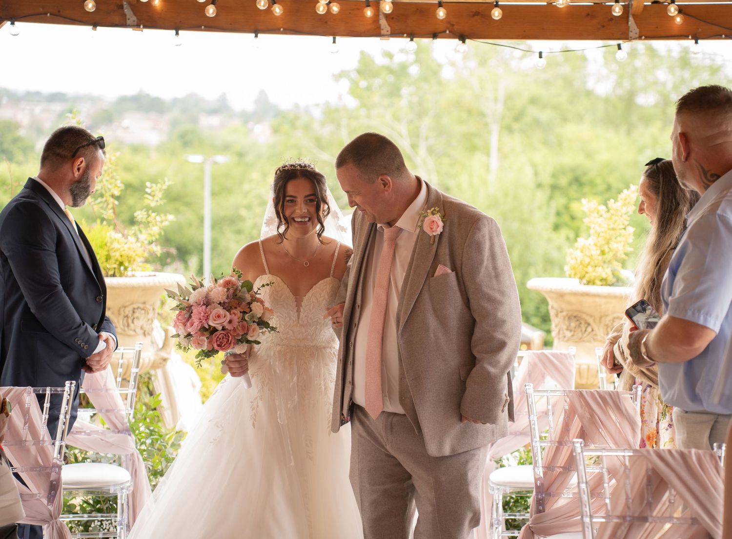 Bride walking down aisle with escort in white ballgown at outdoor wedding ceremony.