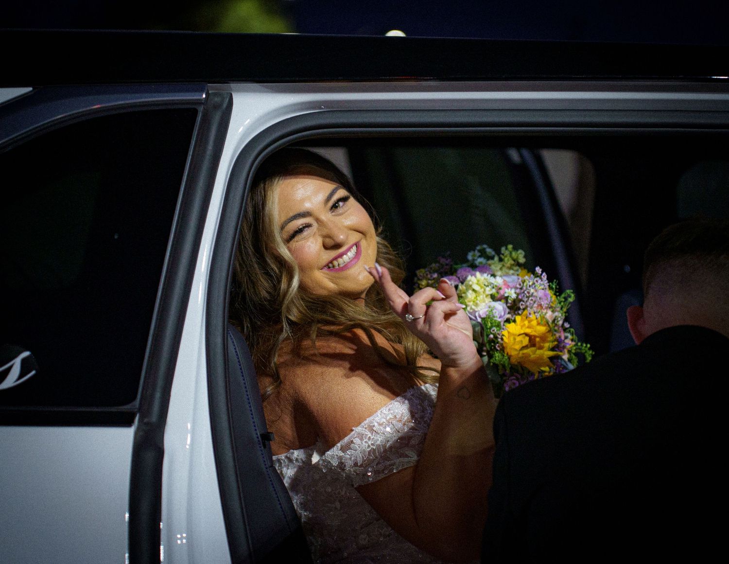 Smiling bride holding colorful bouquet leaning out of white limousine window.