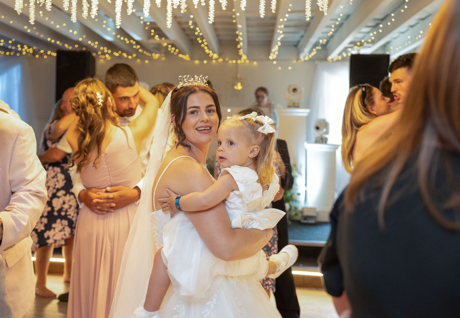 A smiling bride in a tiara holds a flower girl in a white dress at a wedding reception.