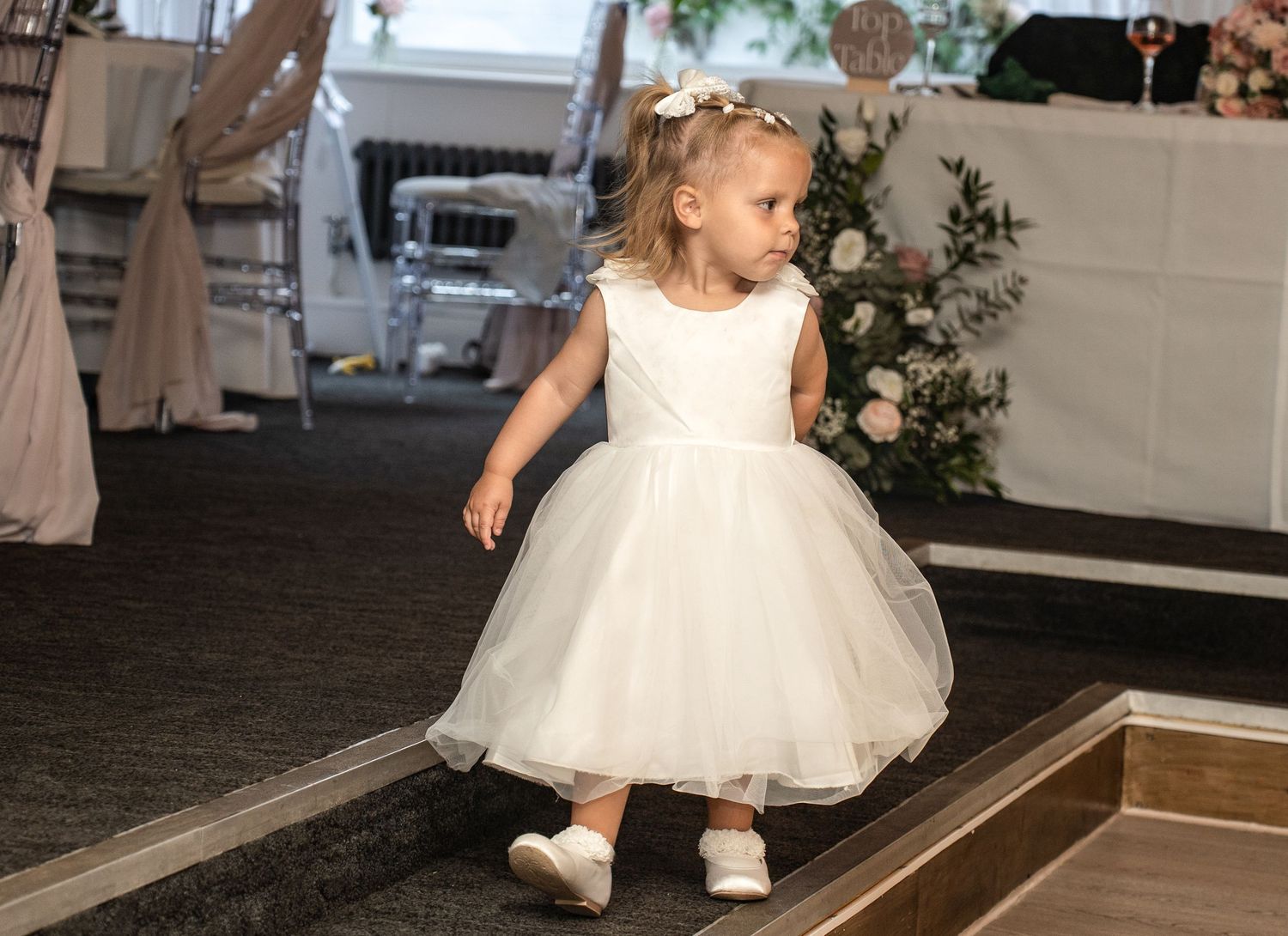 A young flower girl in a white tulle dress walks confidently at a wedding reception venue.