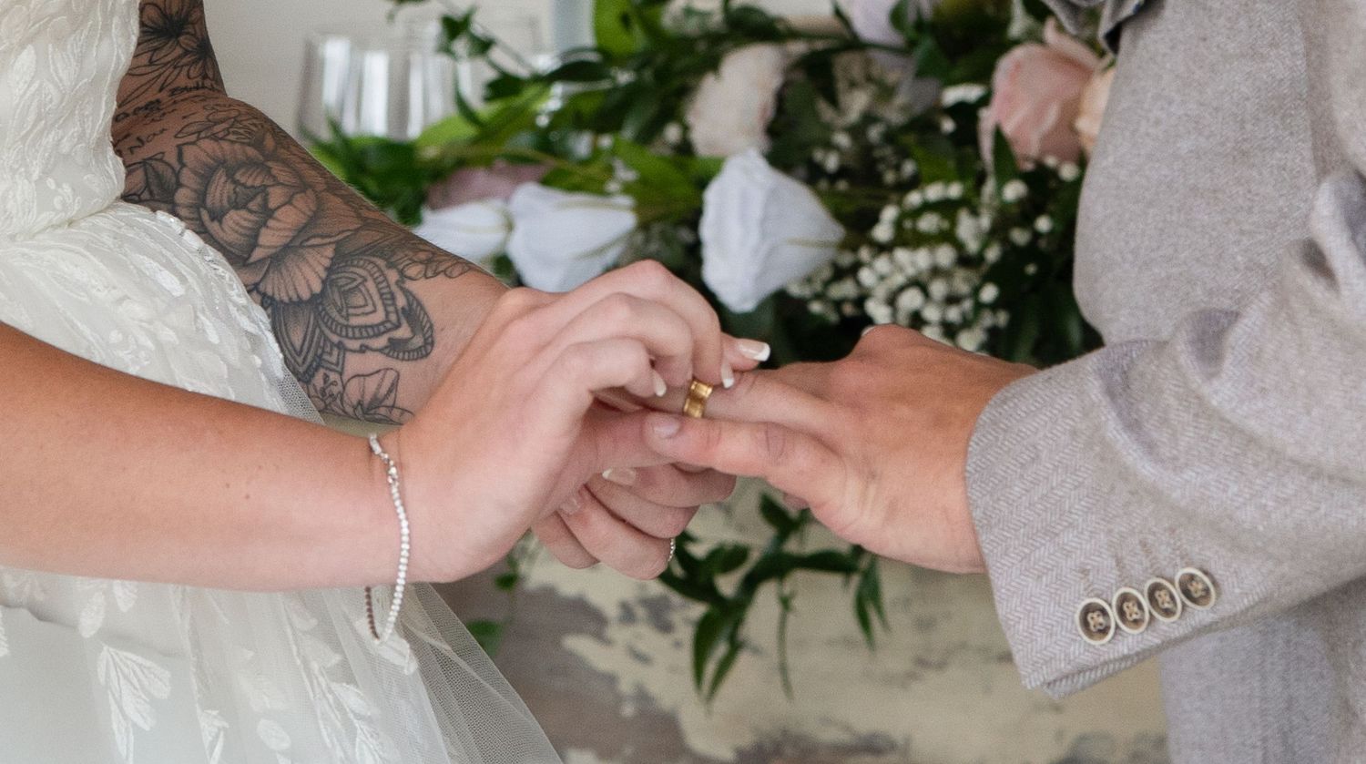 Groom placing wedding ring on brides finger during ceremony, bride has tattoo.