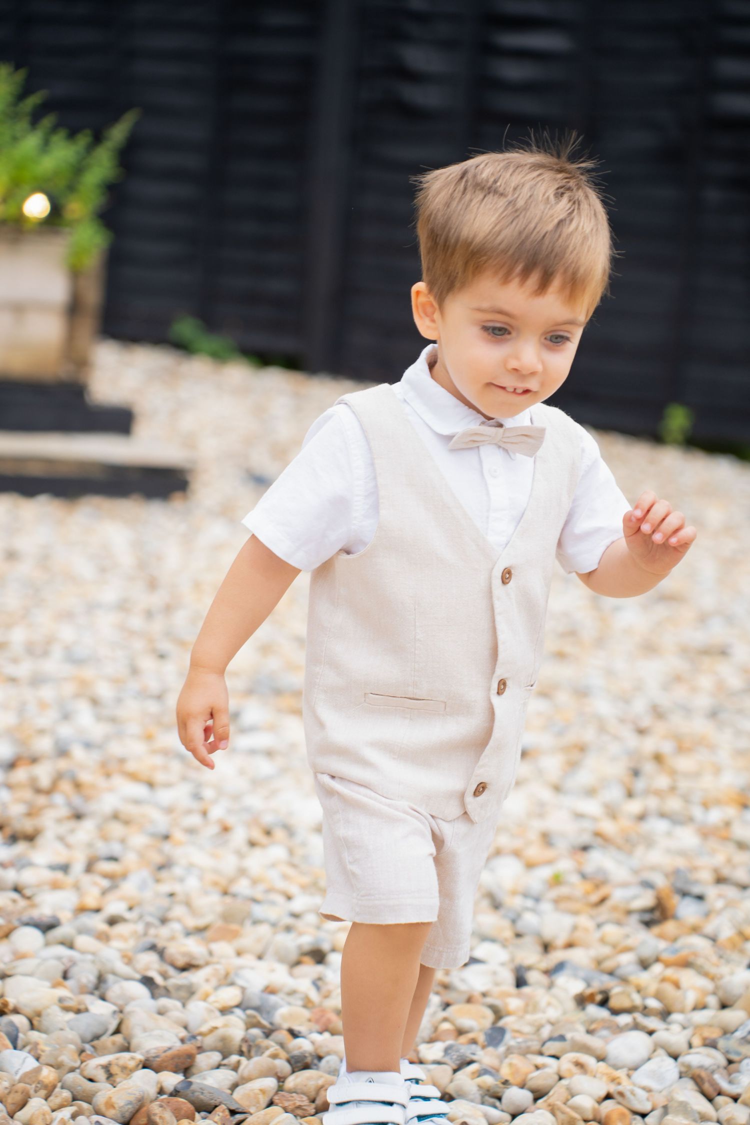 Adorable young ring bearer in beige linen vest and bow tie walking on pebble path.