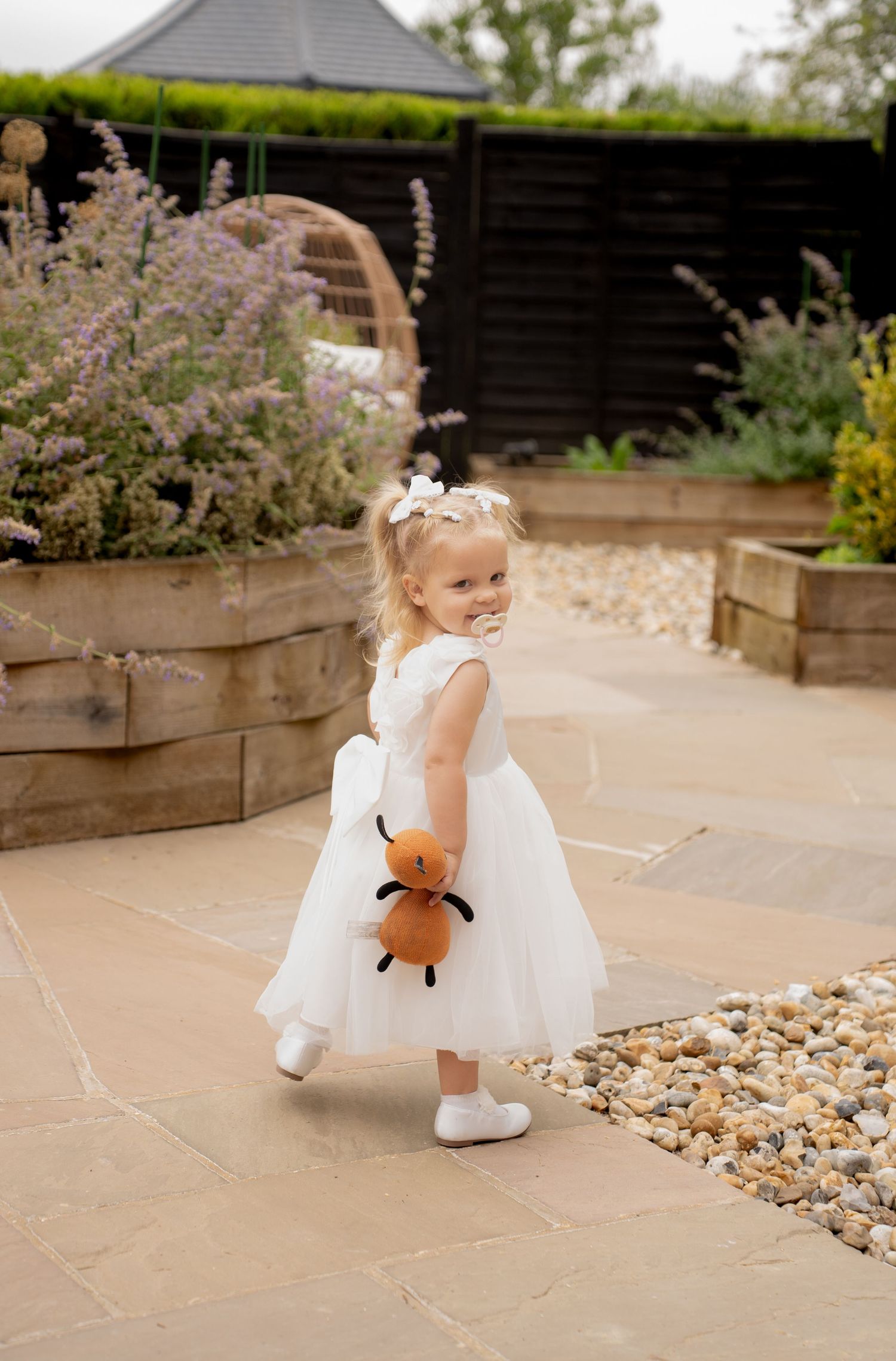 Sweet blonde flower girl in white tulle dress holding stuffed fox toy in garden setting.